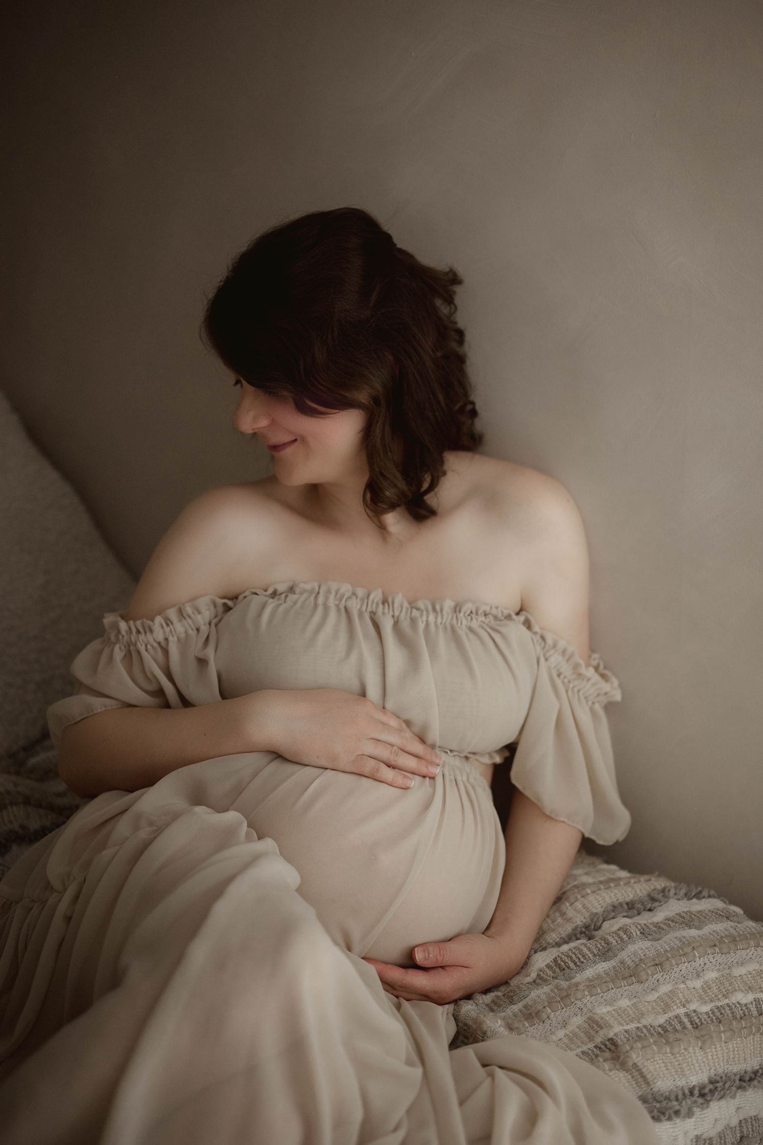 Smiling pregnant woman in beige off-shoulder maternity dress tenderly holding baby bump while seated on bed.