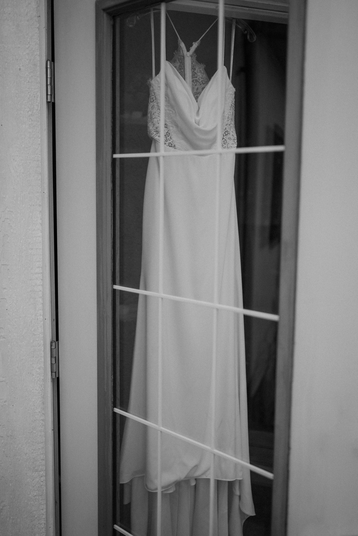 Elegant white wedding dress with lace bodice hanging on a glass door in black and white photo.