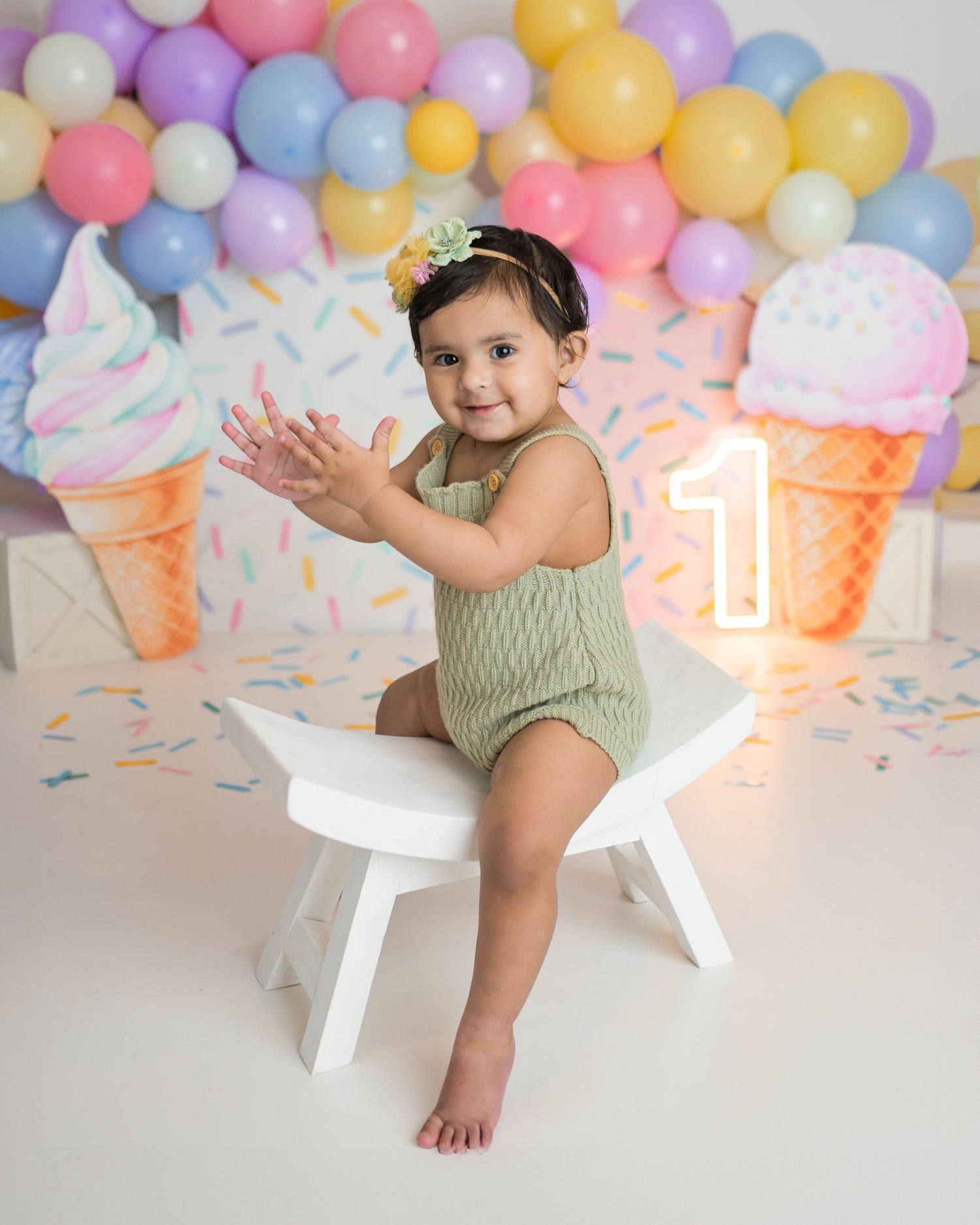 Smiling baby in green romper claps hands at ice cream themed first birthday party with pastel balloon garland.