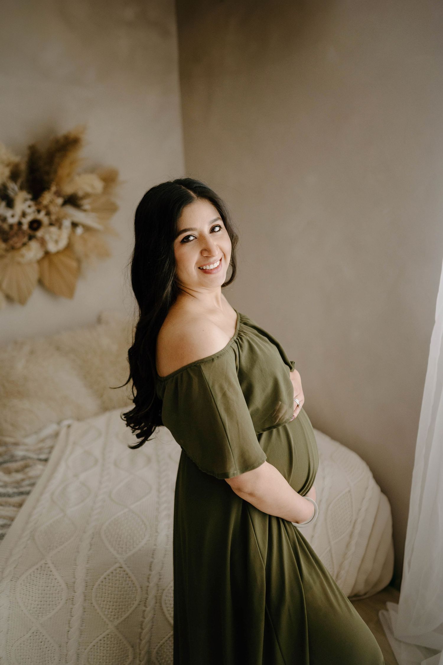 Glowing pregnant woman in olive off-shoulder dress smiling over shoulder in boho bedroom.