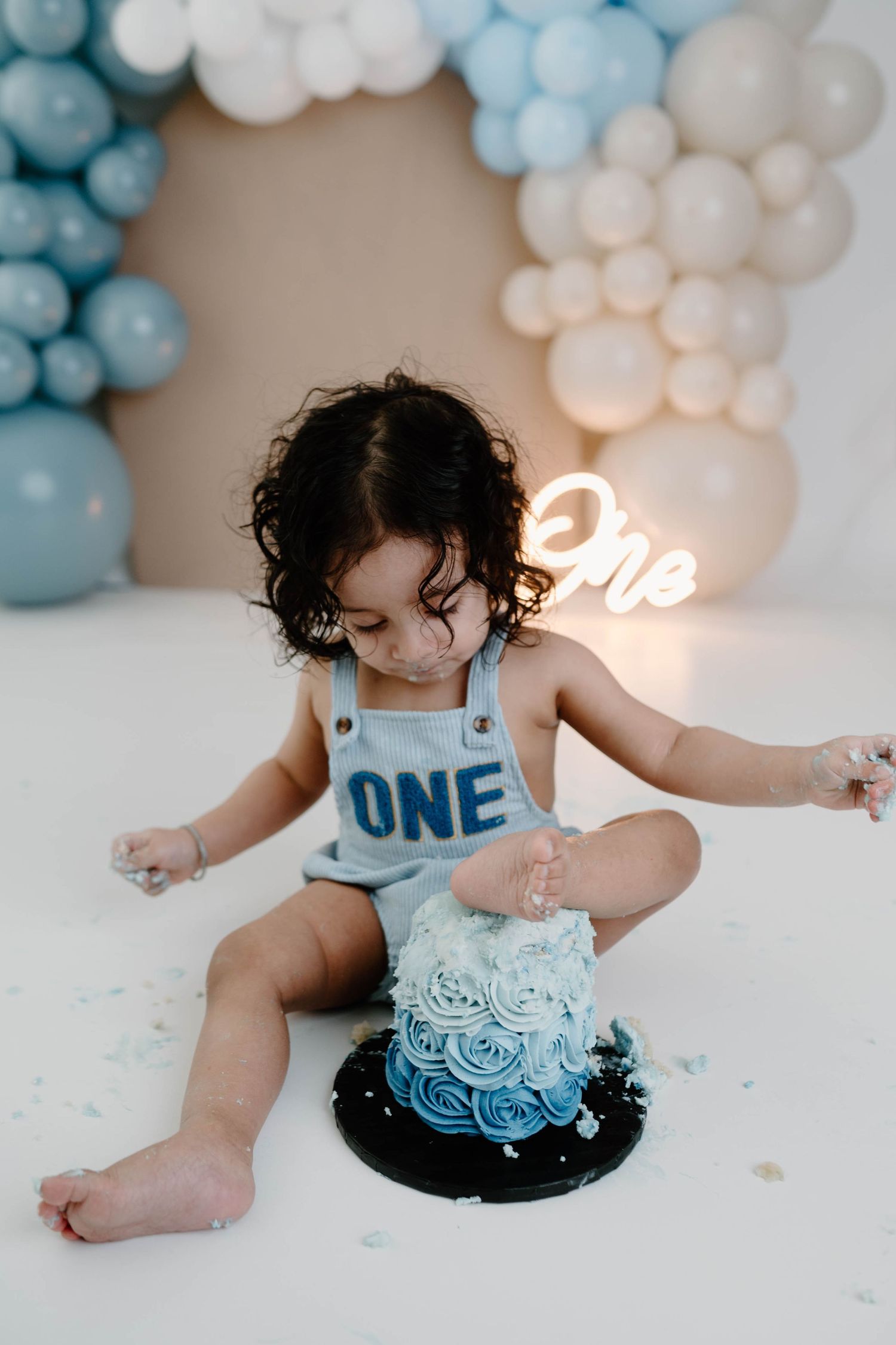 A curly-haired toddler in a blue ONE romper smashes a blue ombre rosette cake with balloon arch backdrop.
