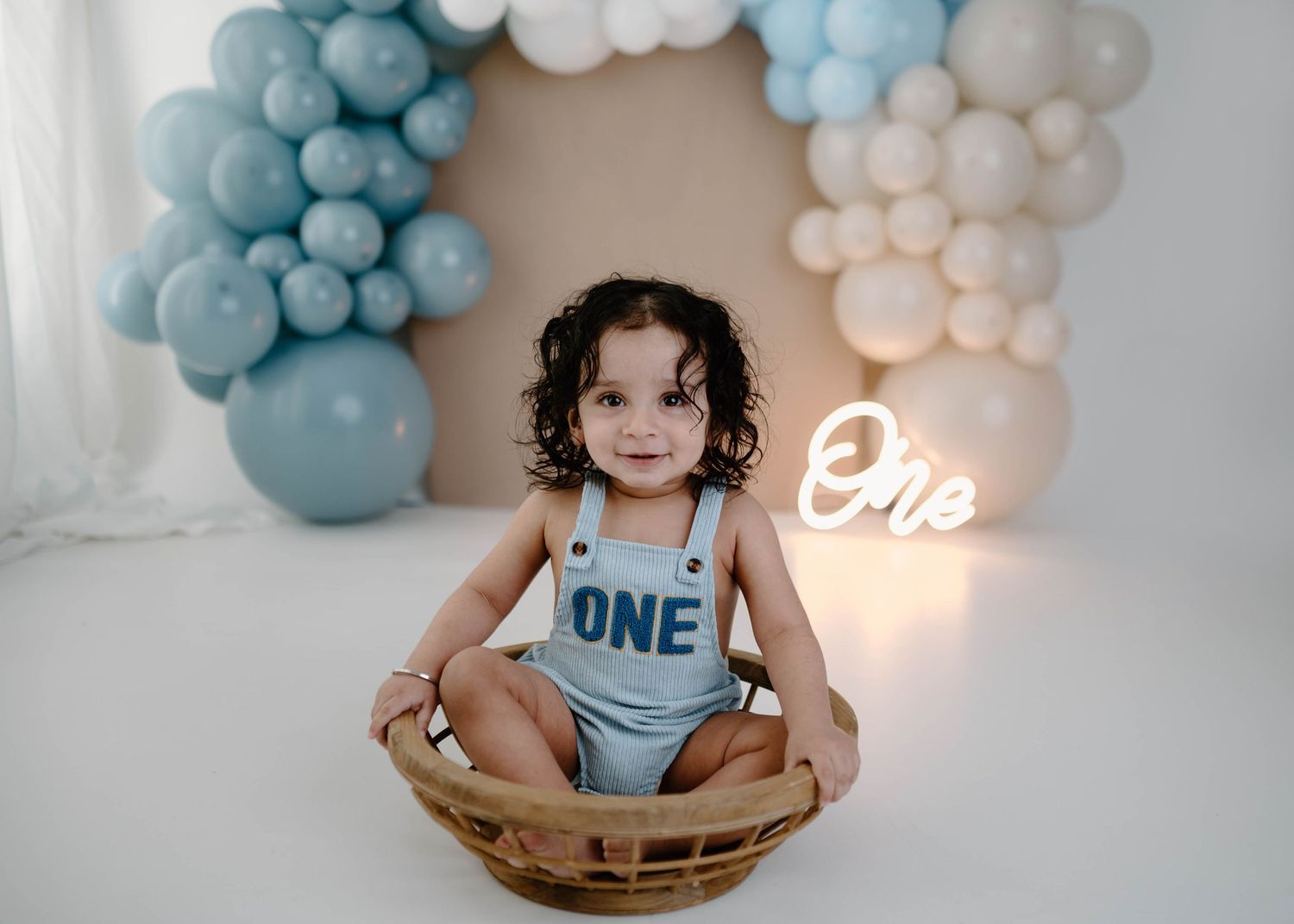 A smiling toddler in a blue ONE romper sits in a wicker basket surrounded by blue and white balloon arch.