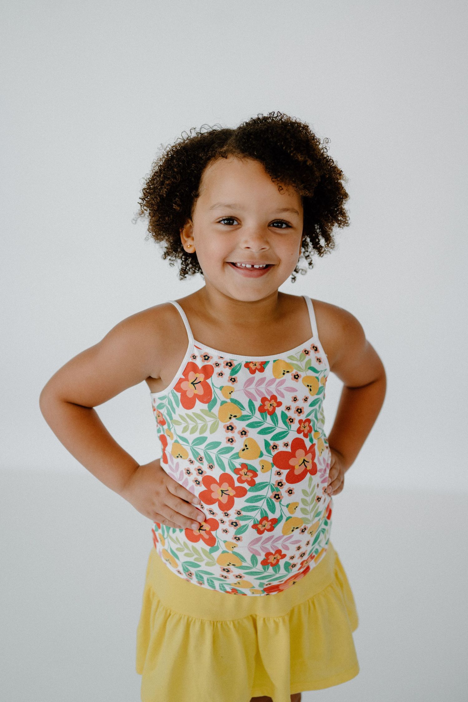 Happy young girl in floral camisole and yellow ruffle skirt posing with hands on hips.