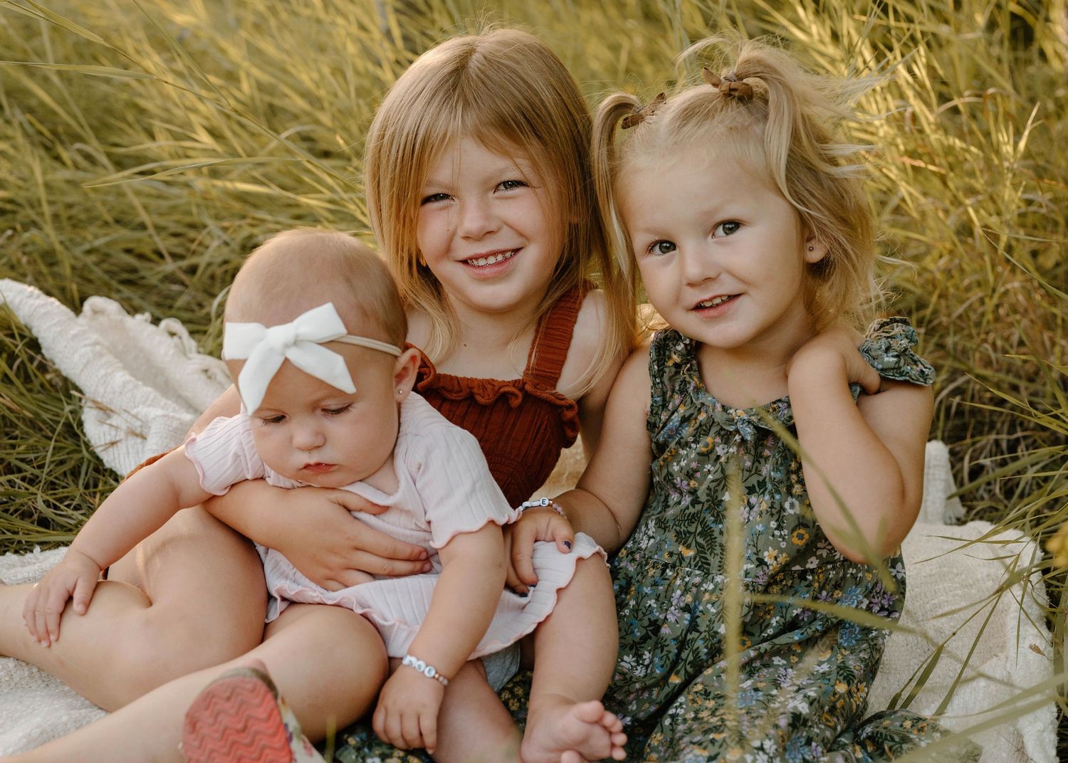 Three young sisters sitting together in golden grass field, two older girls smiling with baby sister.