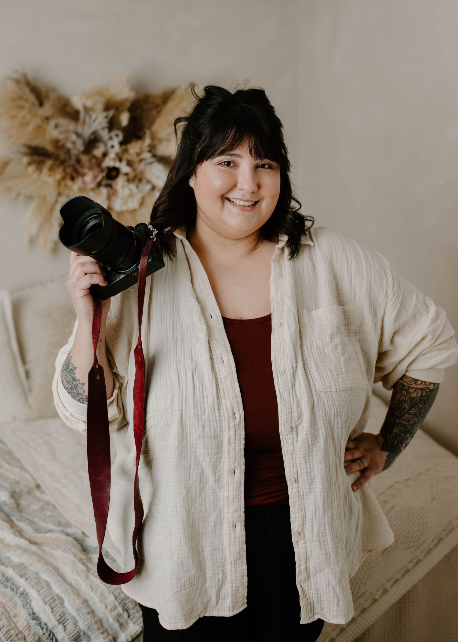 Smiling female photographer holding a DSLR camera with red strap, wearing cream shirt, showing tattoos, in a boho studio.