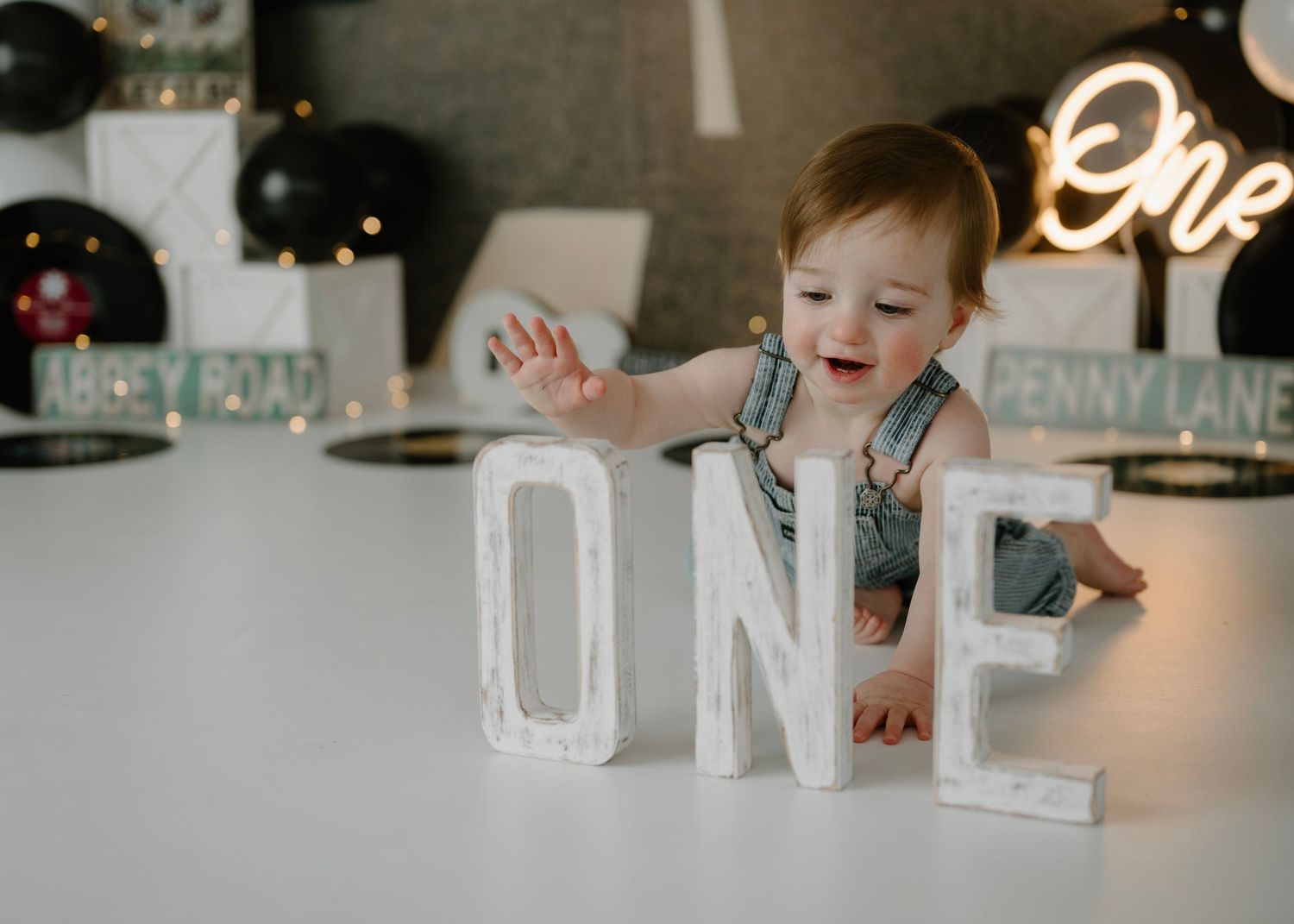 Laughing toddler boy reaches toward large white ONE letter blocks in Beatles-themed first birthday setup.