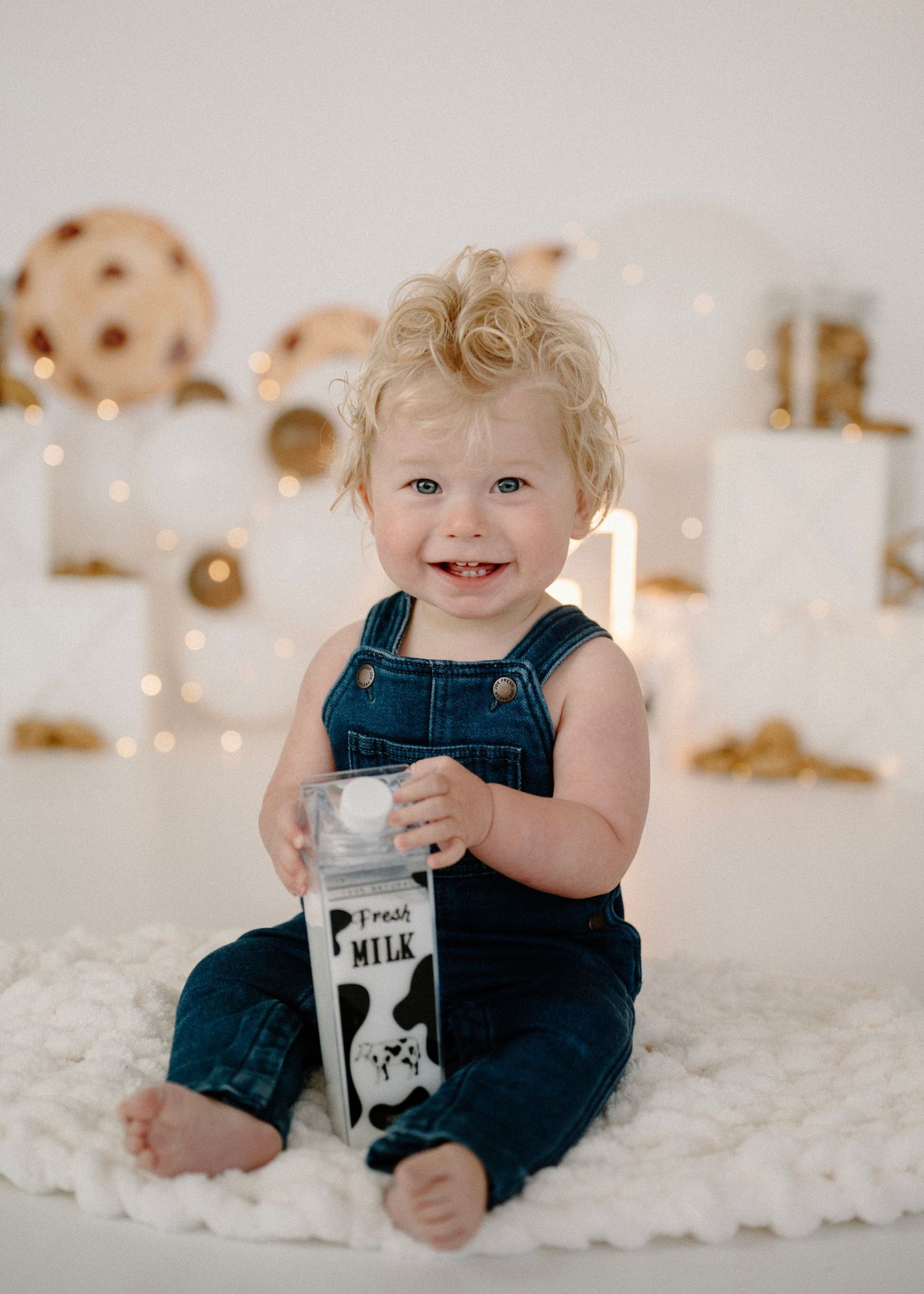 Smiling curly-haired baby in denim overalls holding milk carton with cookies and milk party backdrop.
