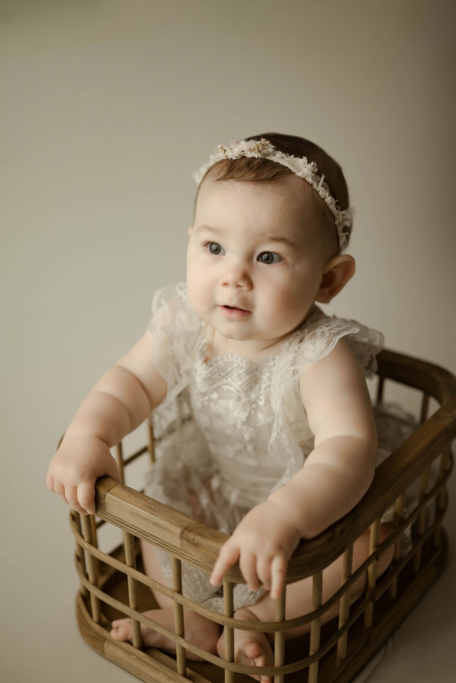 Baby girl in ivory lace romper and floral headband sitting inside rustic wicker basket during studio milestone session.