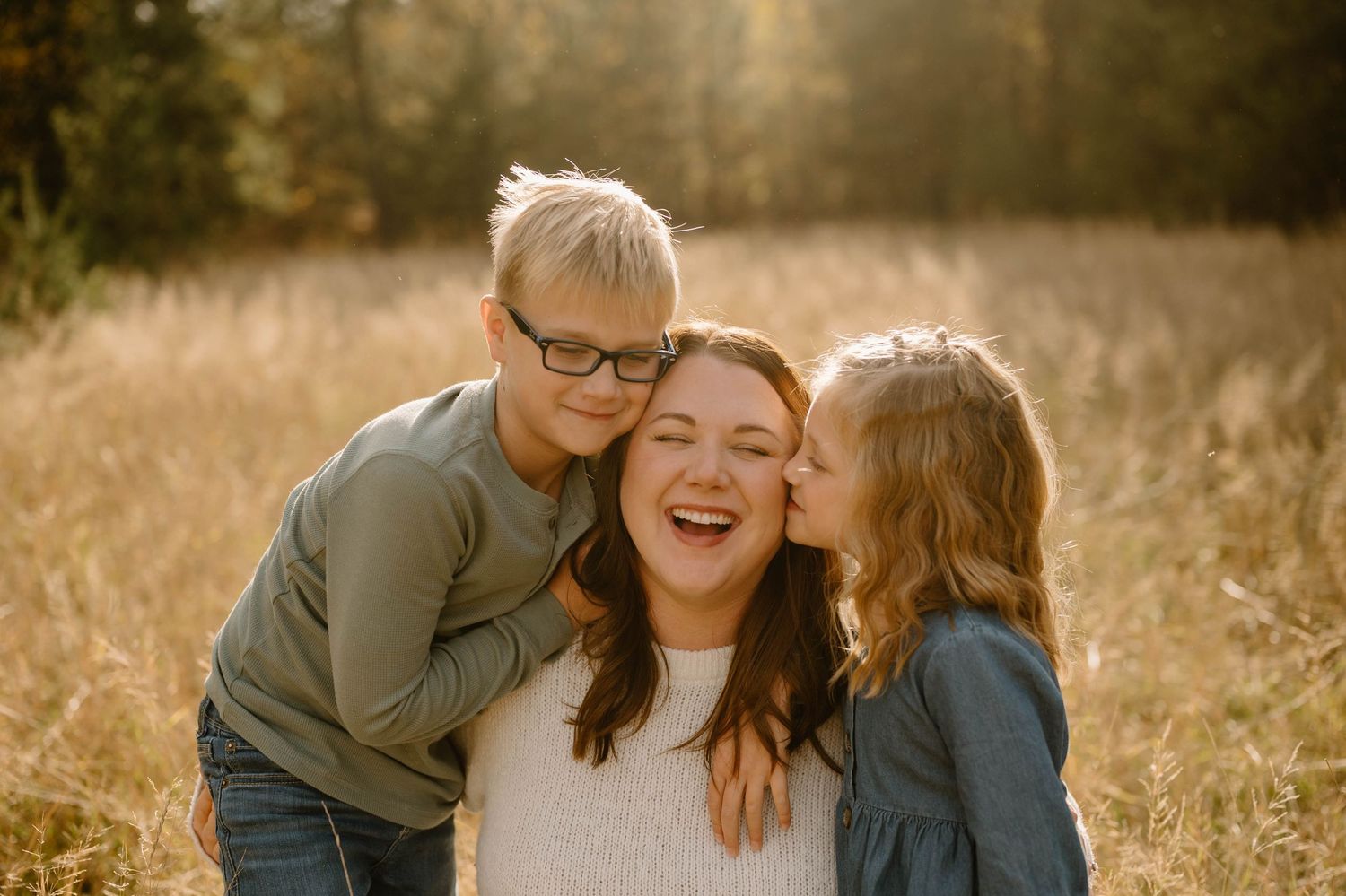 A laughing mother is kissed on both cheeks by her two young children in a field.