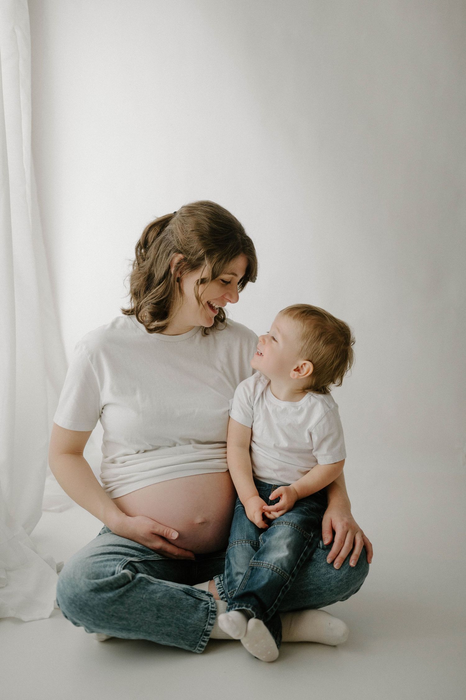 Smiling pregnant woman sits cross-legged with happy toddler boy on white studio floor.