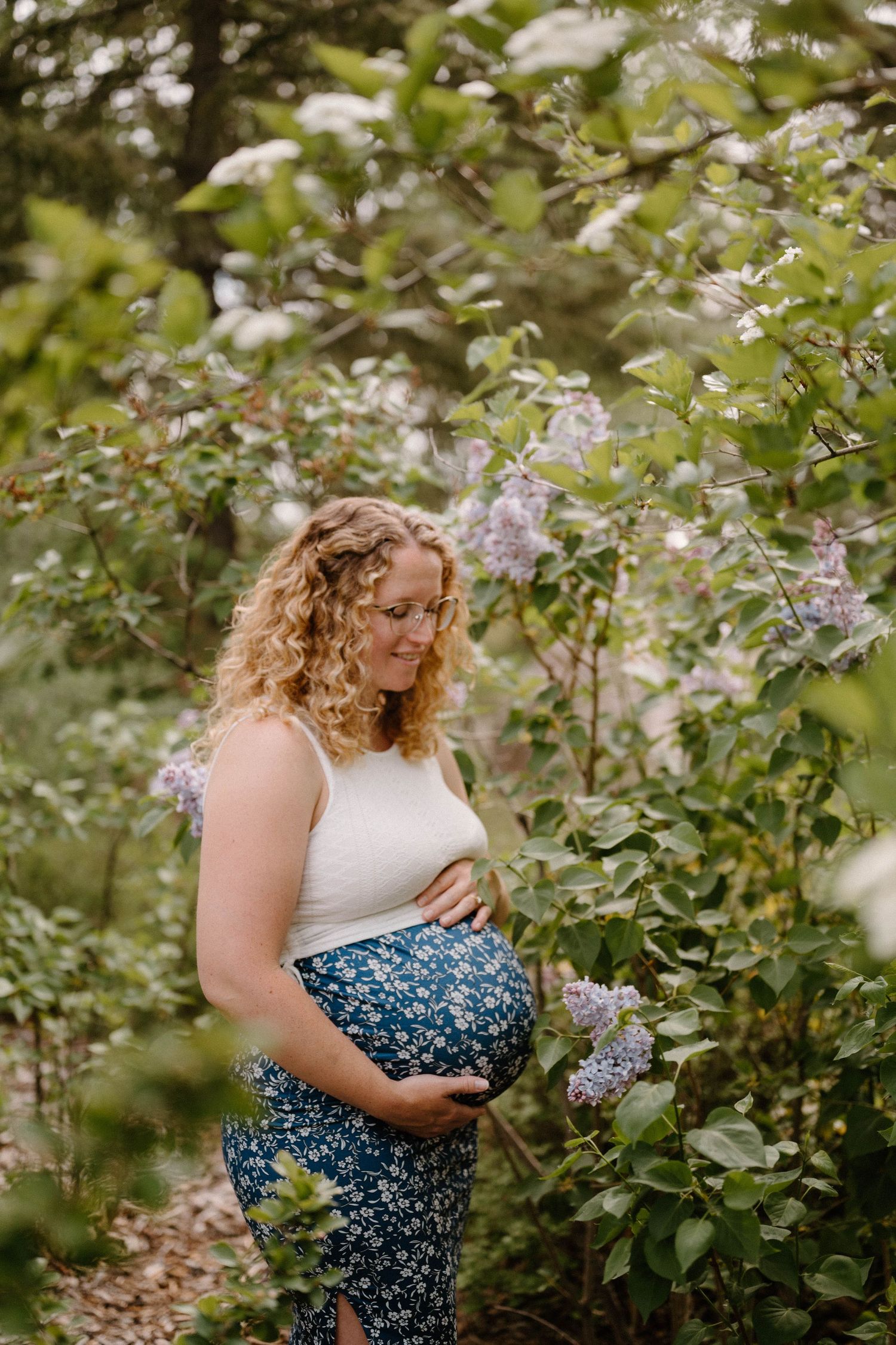 Pregnant woman in floral skirt cradling baby bump among blooming lilac bushes in a lush garden setting.