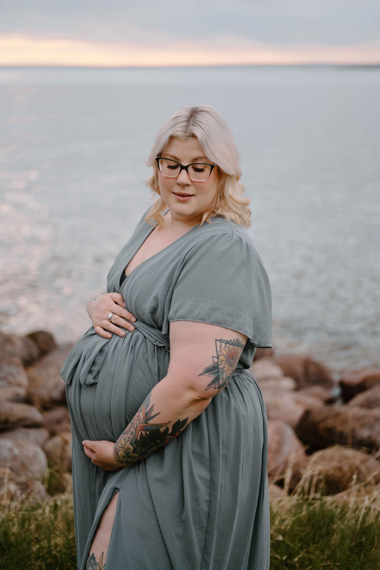 Pregnant woman in teal maxi dress with tattoos cradling baby bump by rocky seaside at sunset.