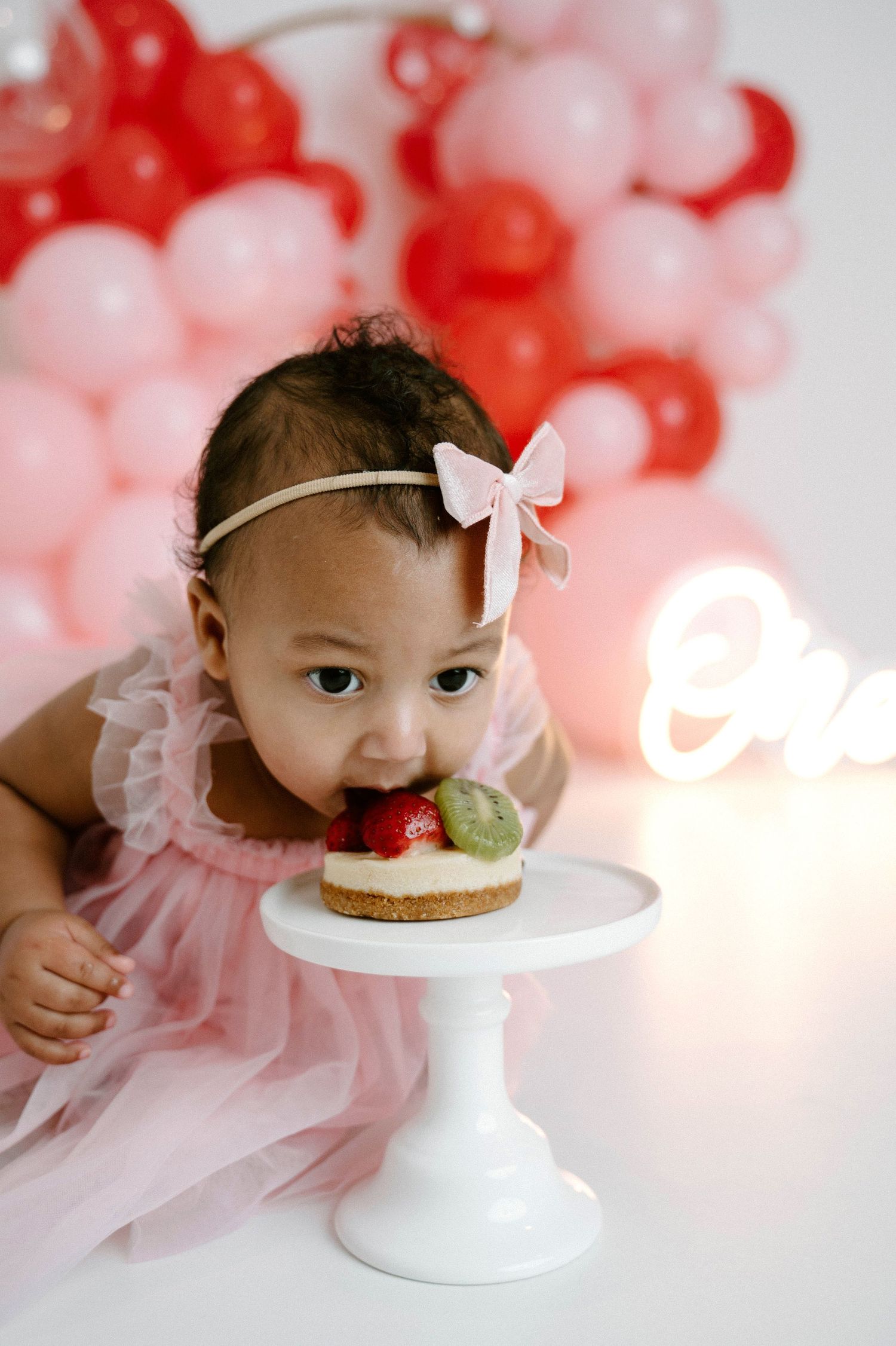 Baby girl in pink dress bites into fruit cake smash on white pedestal with balloon backdrop in Leduc.