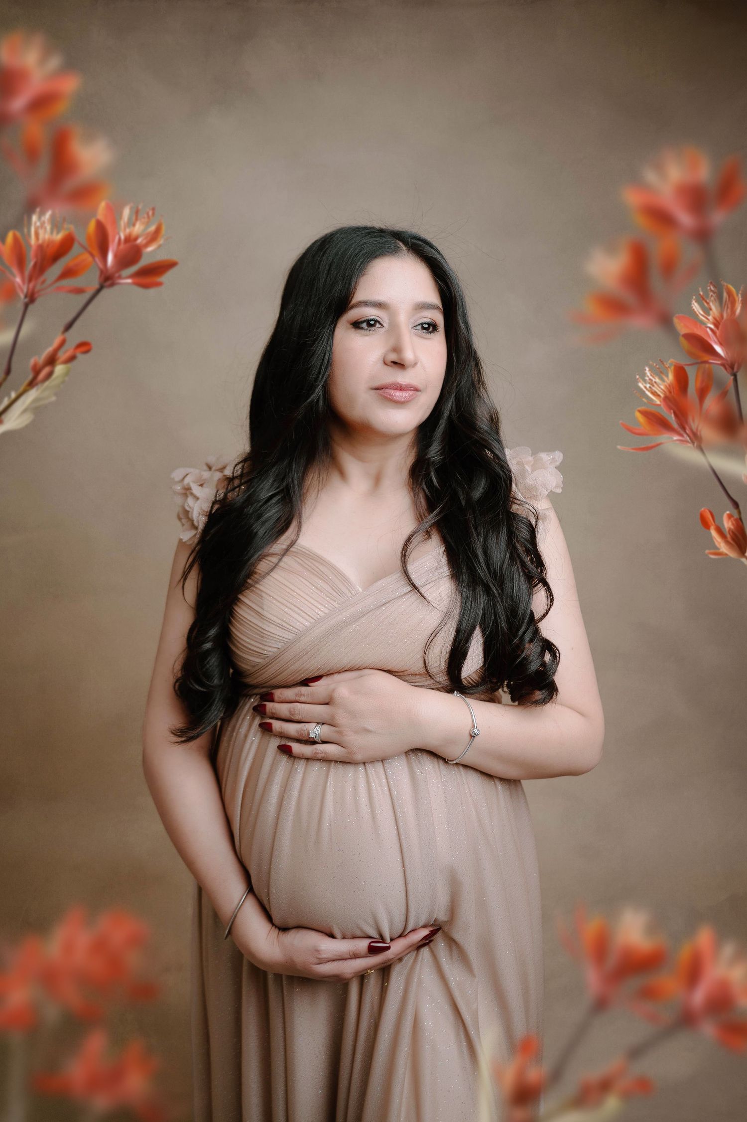 A pregnant woman in a beige gown holds her belly surrounded by orange floral branches in a studio.