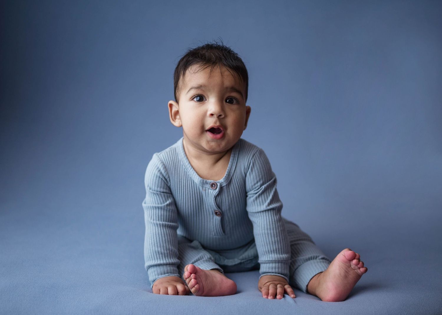 Curious baby in a light blue ribbed romper sitting upright on a blue studio floor with a surprised expression.