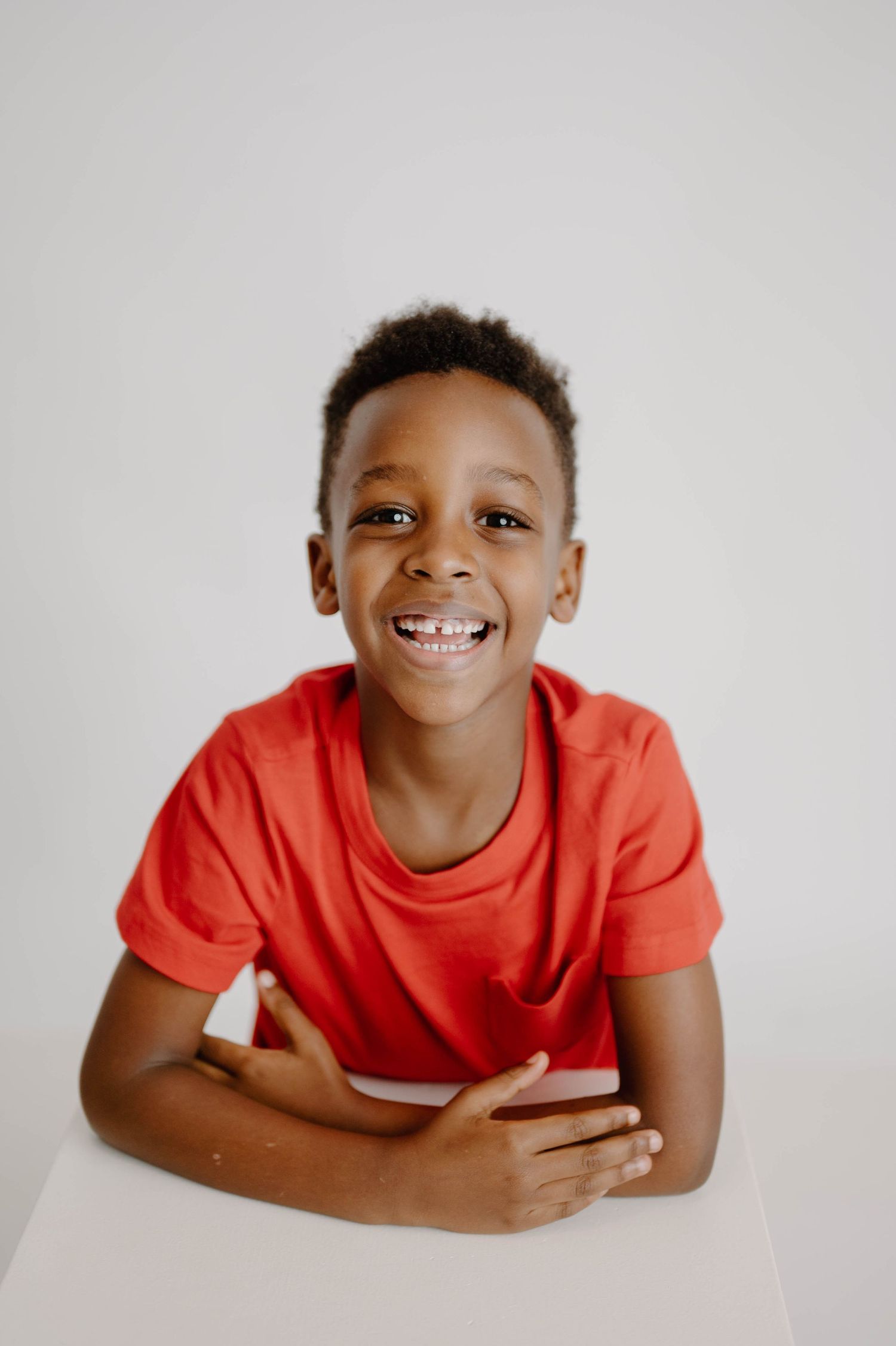 Laughing young Black boy in a red t-shirt sitting at a white table with arms crossed.