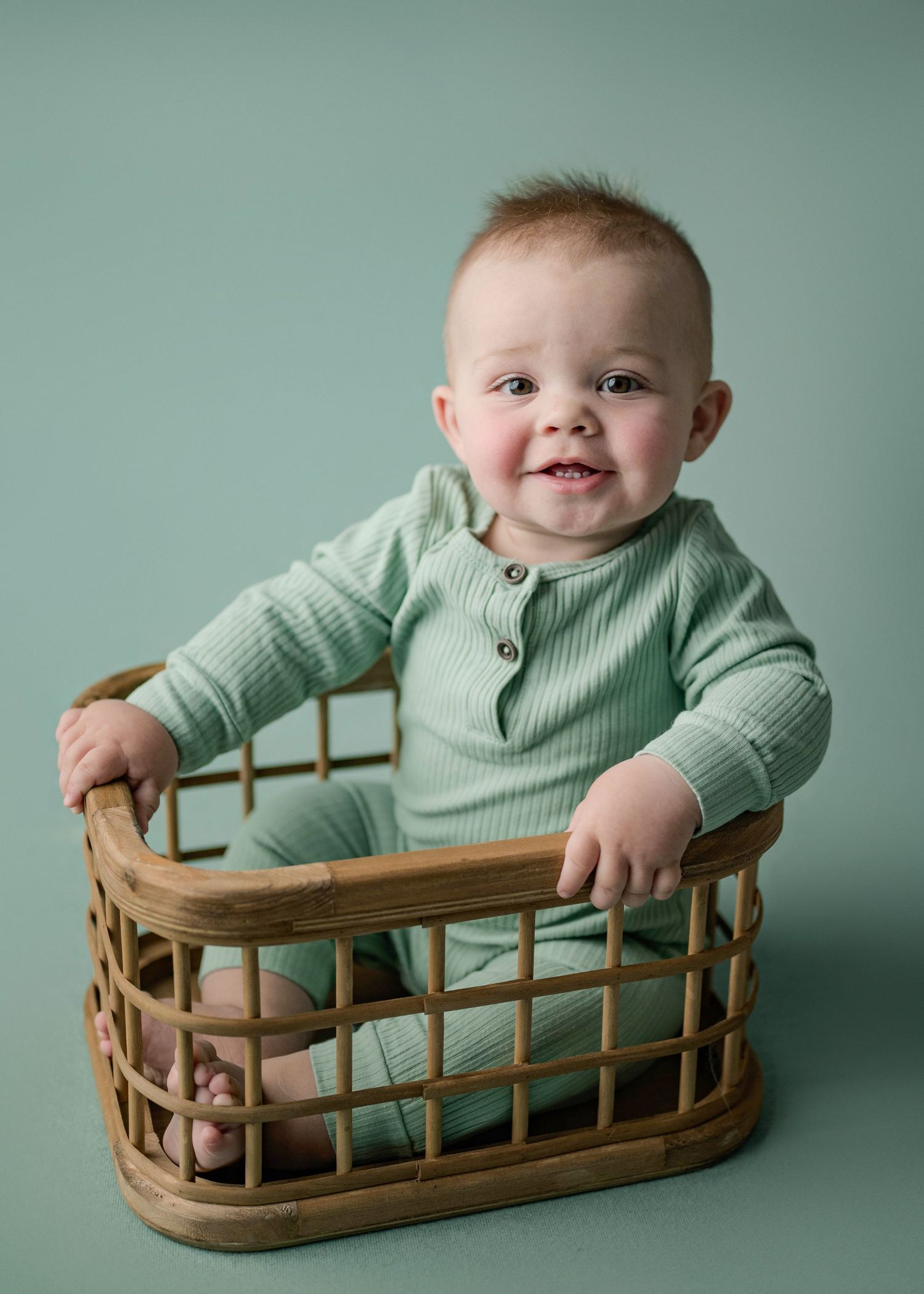 Happy baby boy in green ribbed romper sitting inside a wicker basket on teal background.