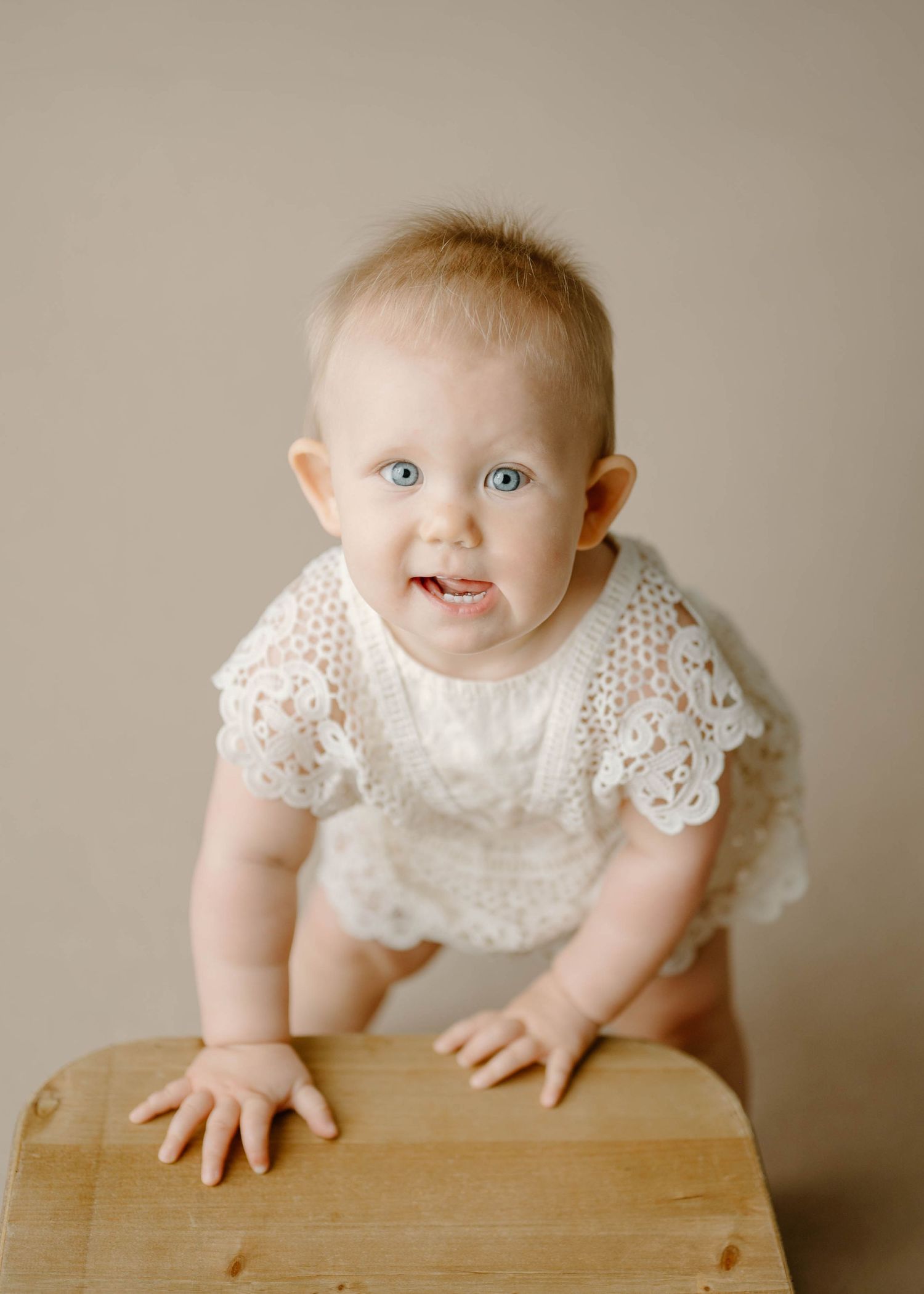 Smiling baby girl with blue eyes in a white lace romper leaning on a wooden stool in a studio.