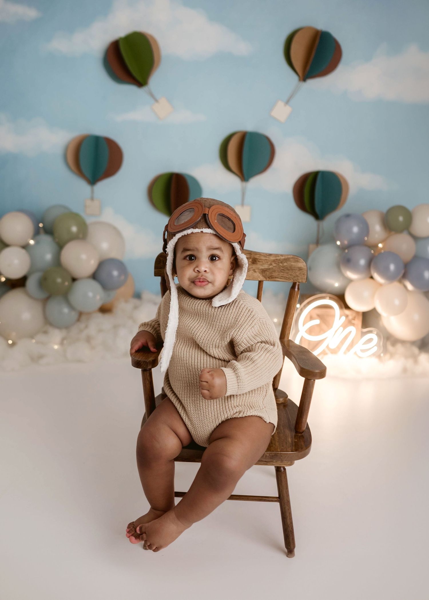 Adorable baby in aviator hat and tan romper sitting in a wooden chair at a hot air balloon first birthday session.