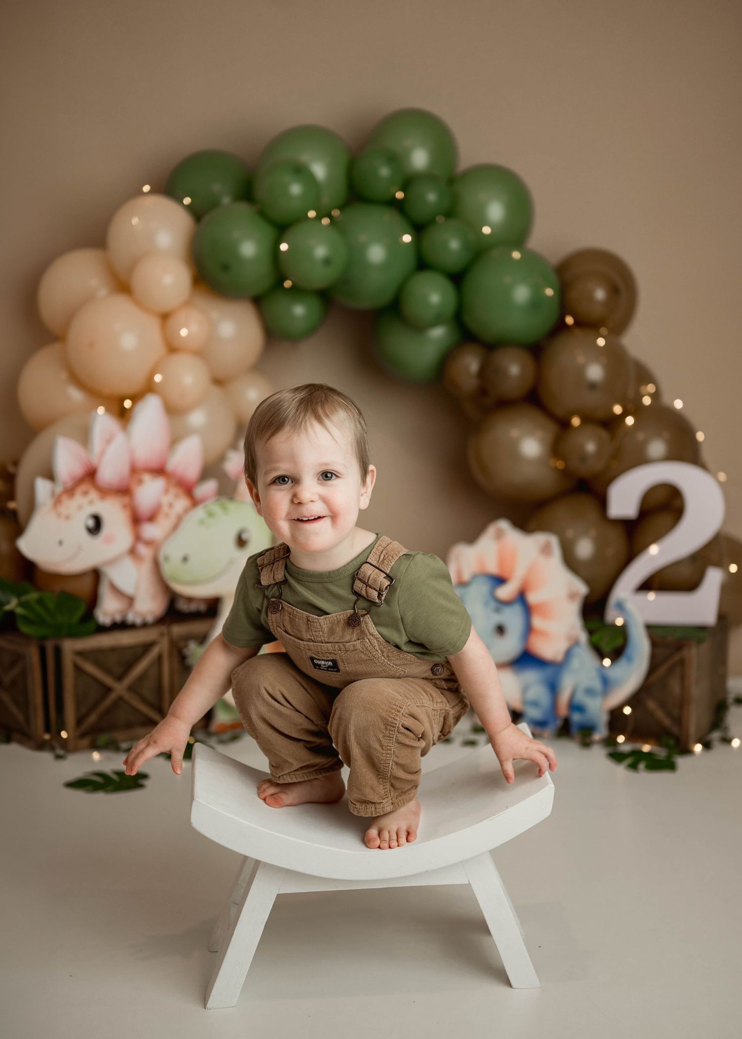 Smiling toddler boy in green shirt and brown overalls sits on white stool at dinosaur-themed 2nd birthday party.