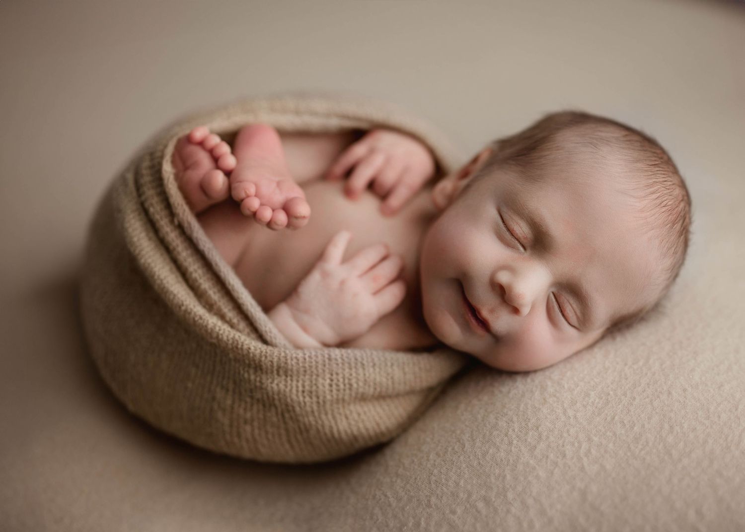 Adorable smiling newborn wrapped in tan knit swaddle showing tiny feet and hands on beige backdrop.