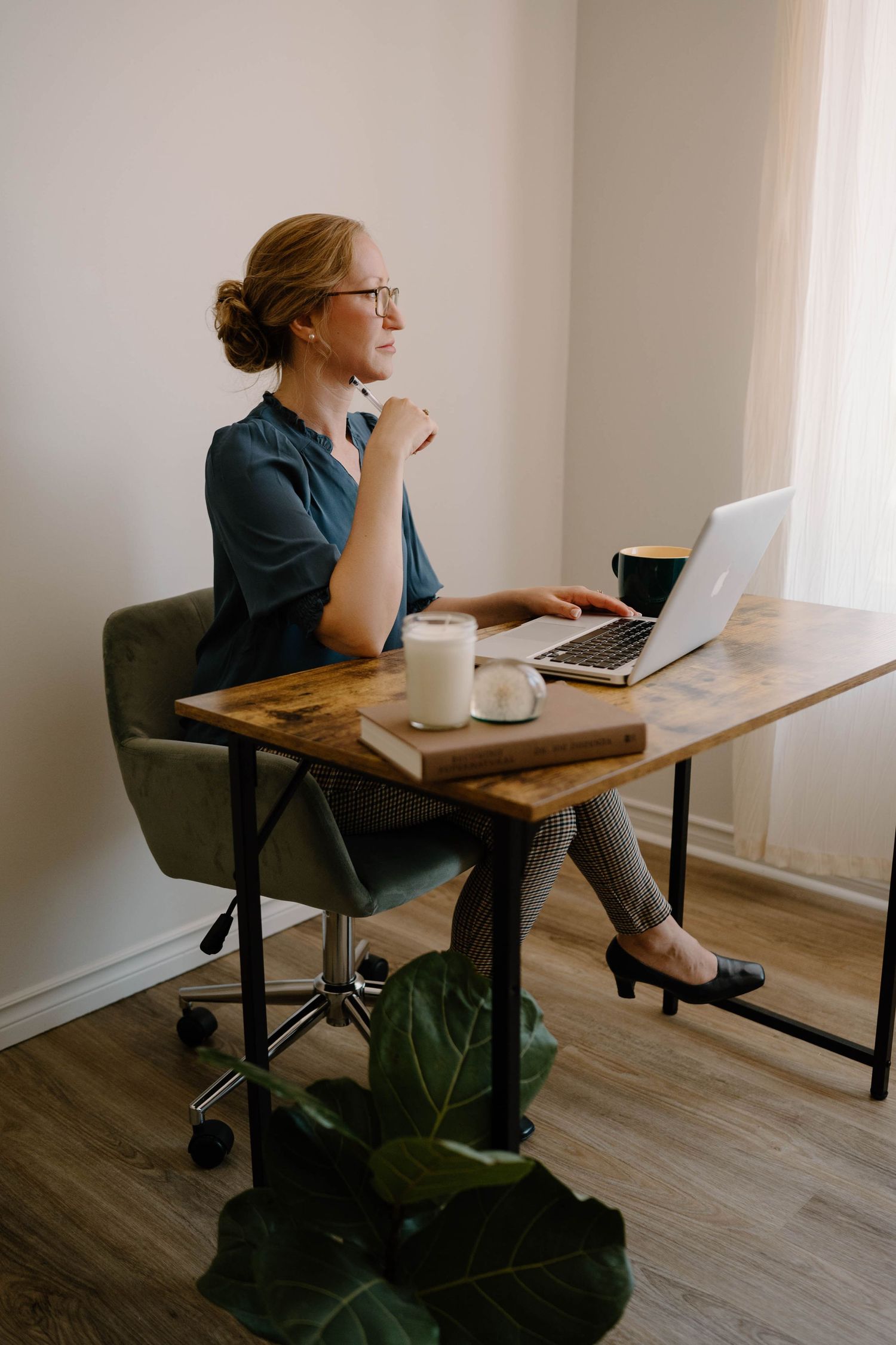 Professional woman in teal blouse sitting at wooden desk with laptop, gazing thoughtfully out window.