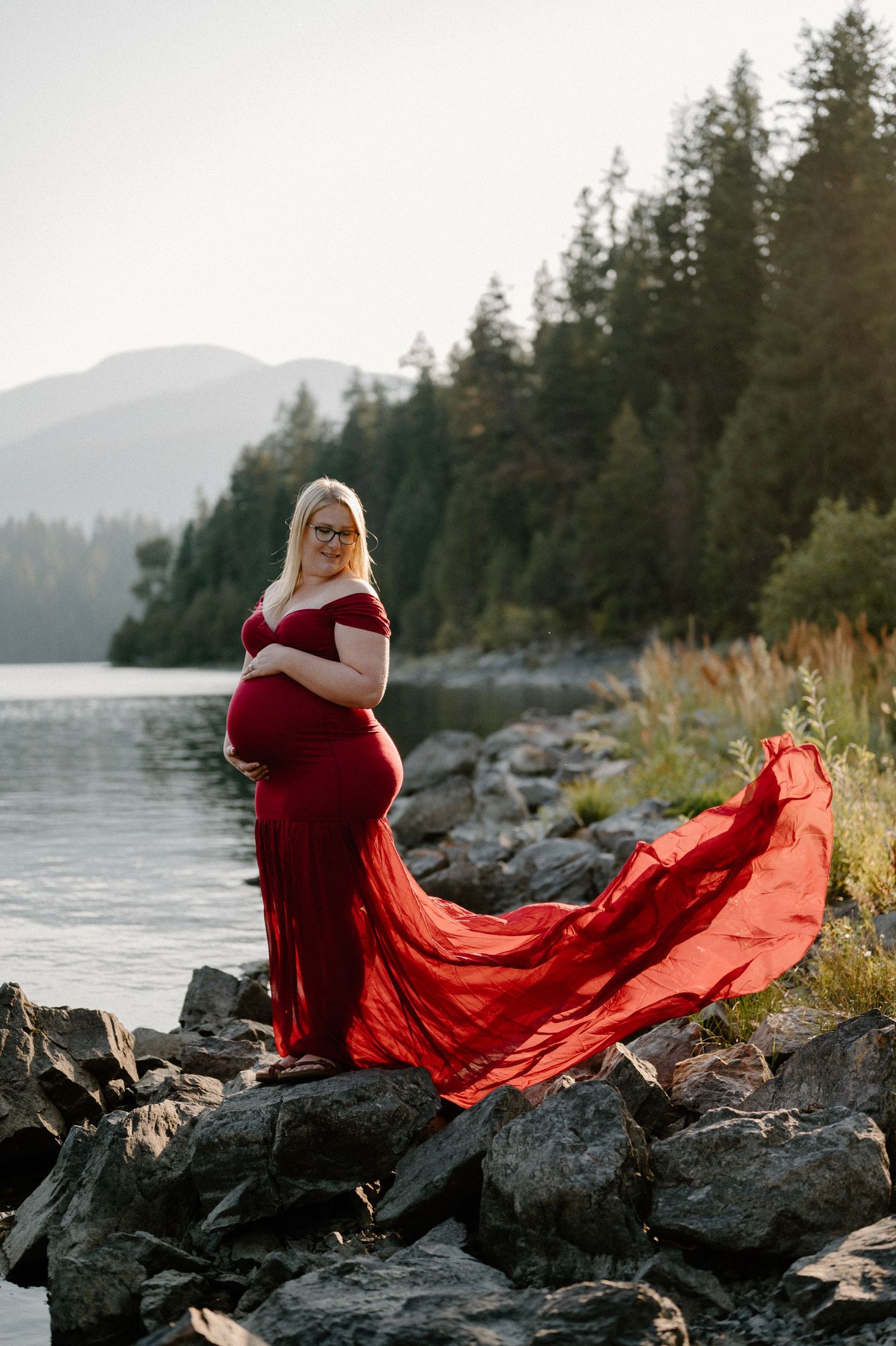 Pregnant woman in flowing red gown standing on rocky lakeshore with mountain backdrop.