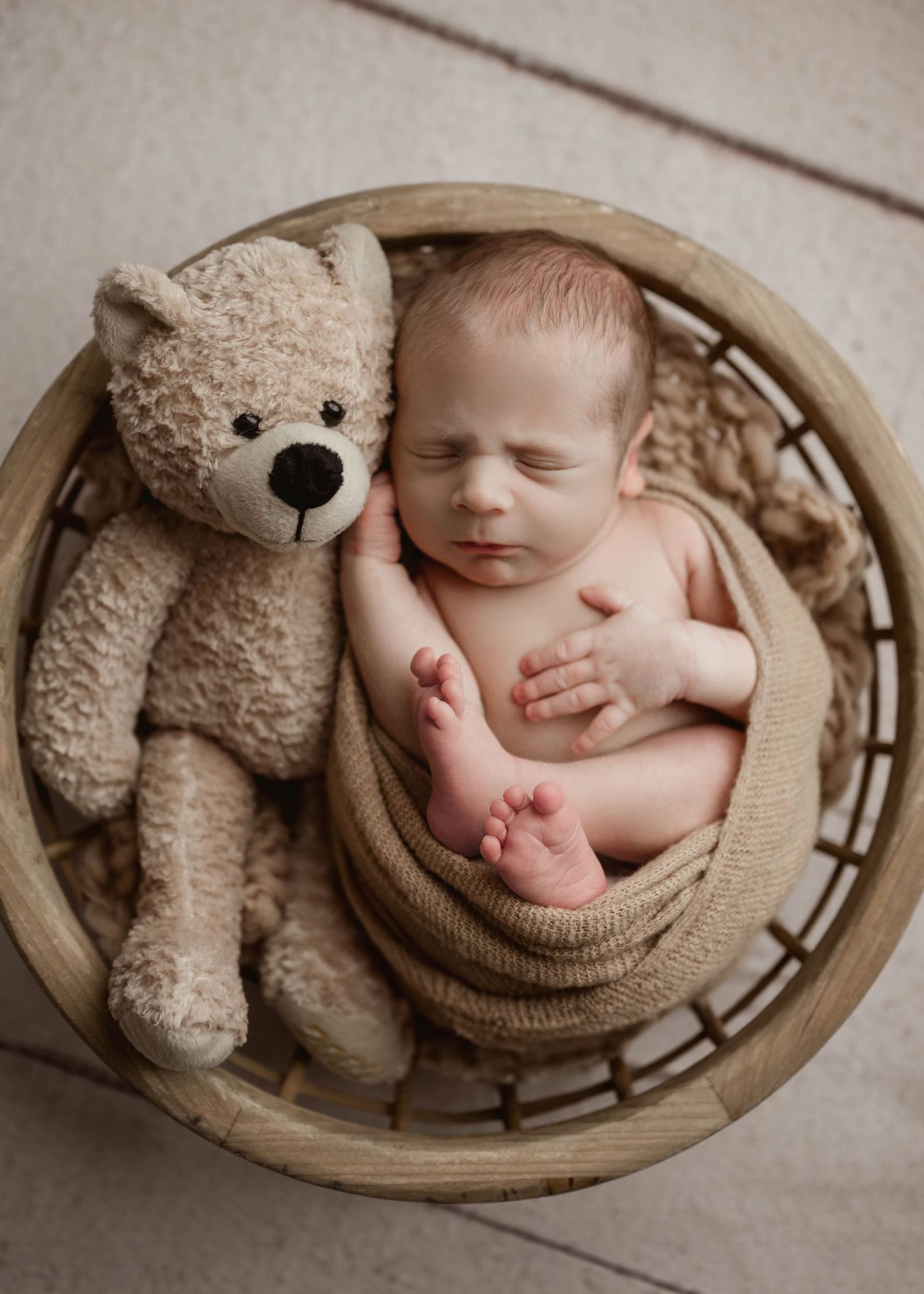 Newborn baby sleeping in wooden basket beside a soft tan teddy bear wrapped in beige knit.