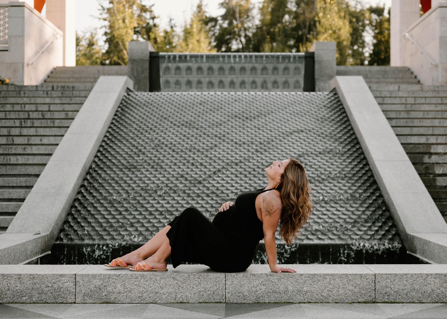 Pregnant woman in a black dress posing gracefully on stone steps beside a waterfall feature.