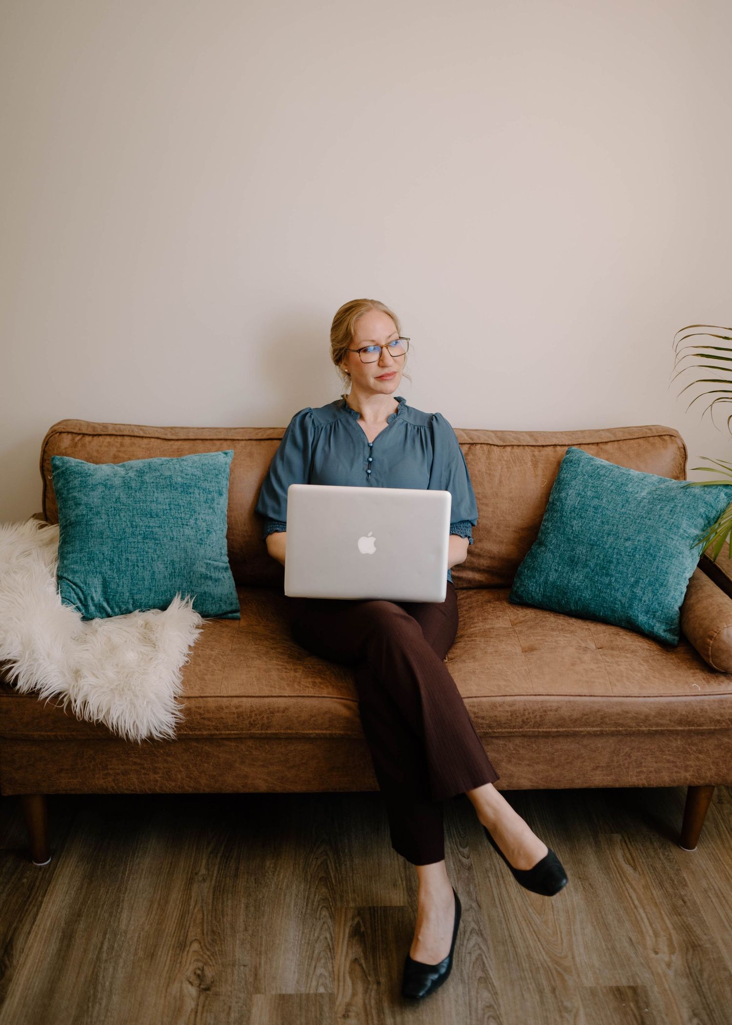 Professional blonde woman in teal top sitting on brown leather sofa working on MacBook laptop.