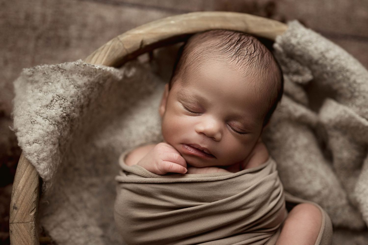 Newborn baby wrapped in beige swaddle sleeps peacefully in a rustic wooden basket with soft textured blanket.