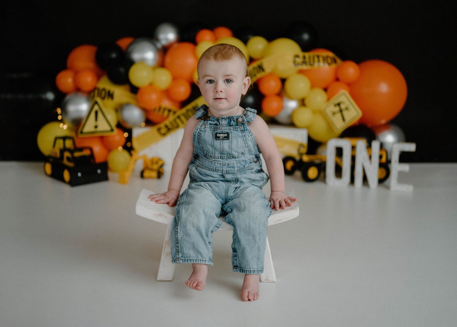 Baby boy in denim overalls sits on white box before construction-themed first birthday balloon backdrop.