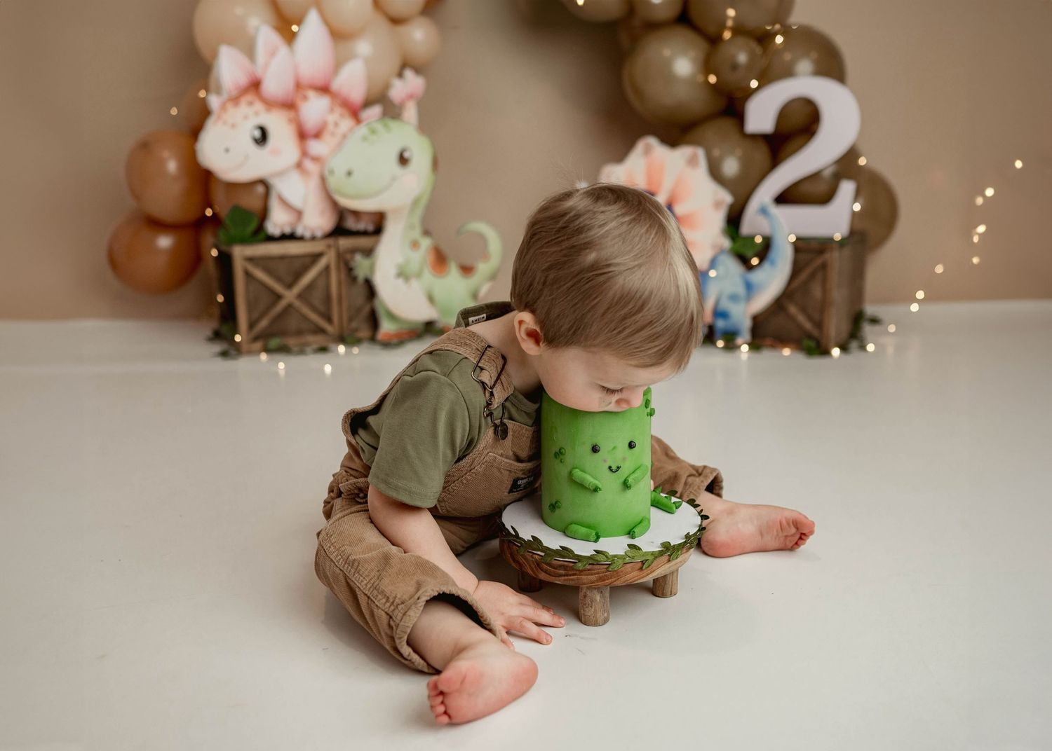 Toddler boy in brown overalls leans over a green dinosaur-decorated birthday cake on white floor.