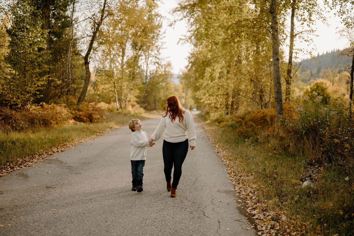 Mother and young child walking hand in hand down a tree-lined autumn road.