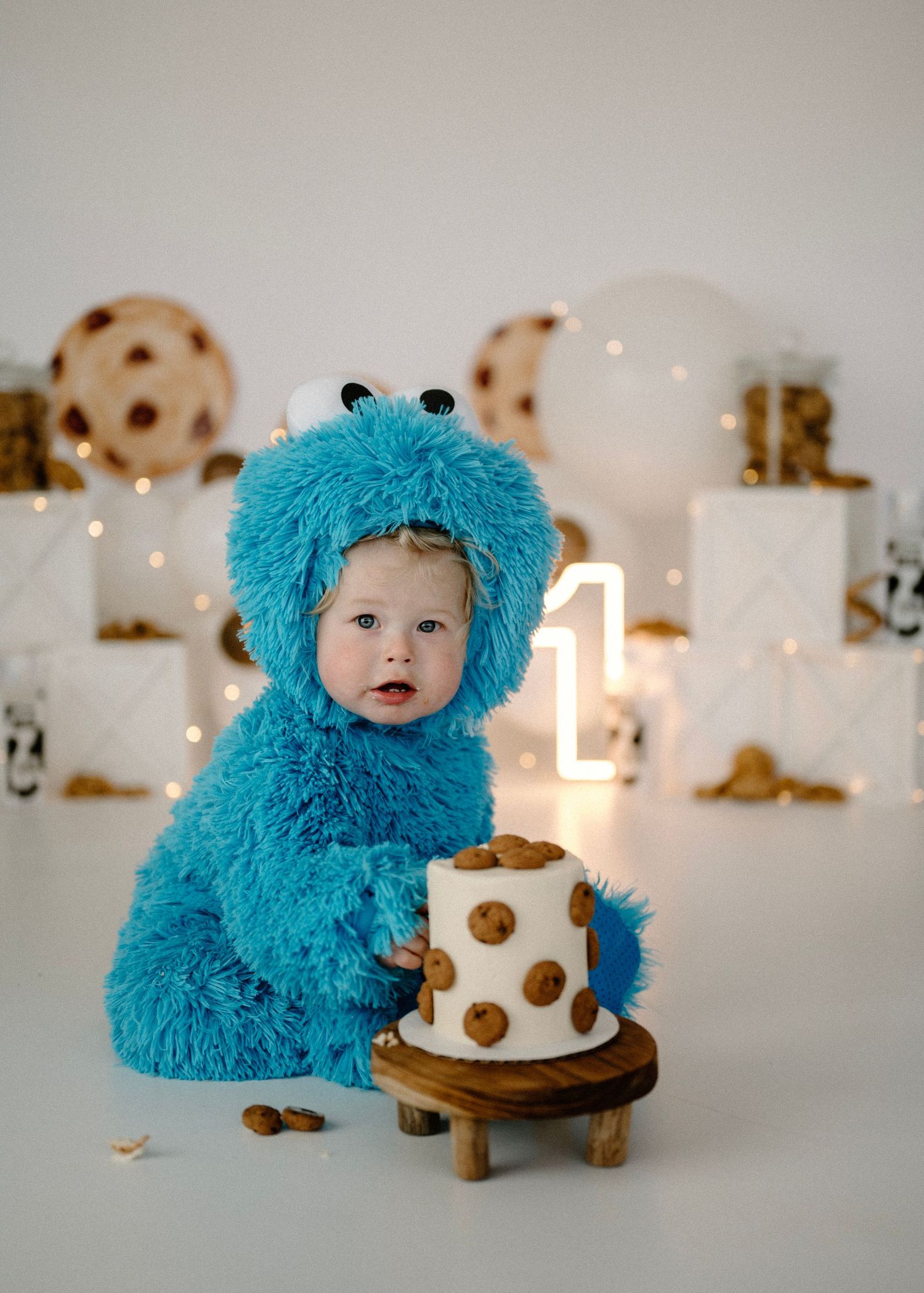 Baby in Cookie Monster costume sitting with chocolate chip cookie cake on wooden stand.