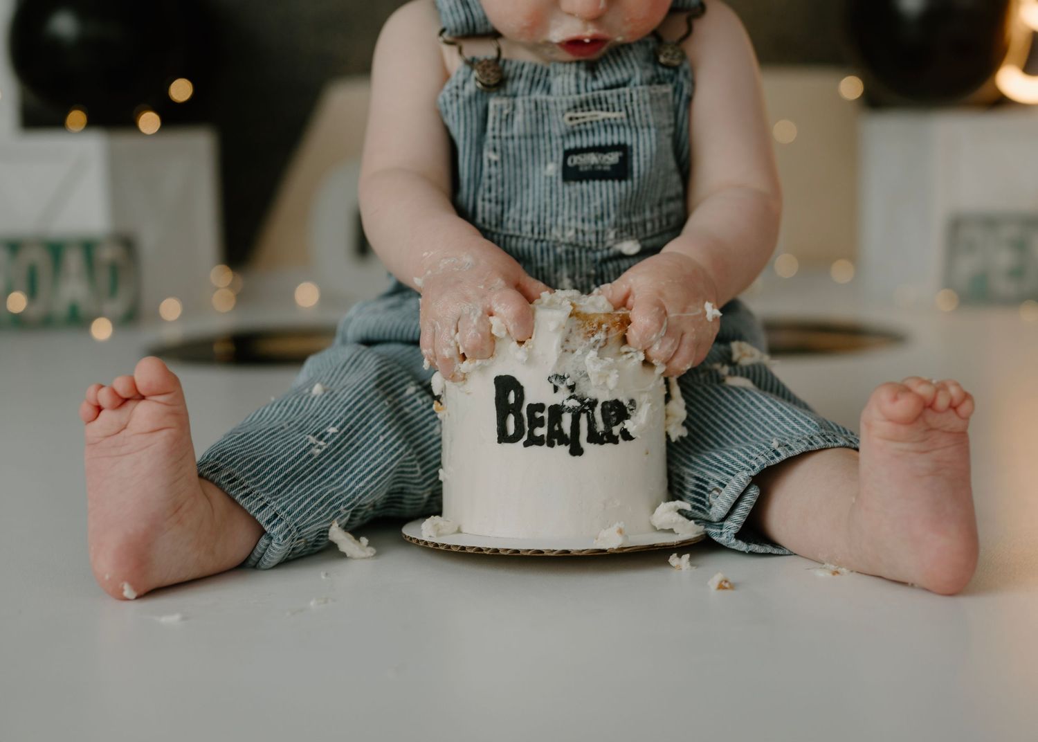 Baby in denim overalls smashes white Beatles-themed first birthday cake with bare feet visible on floor.