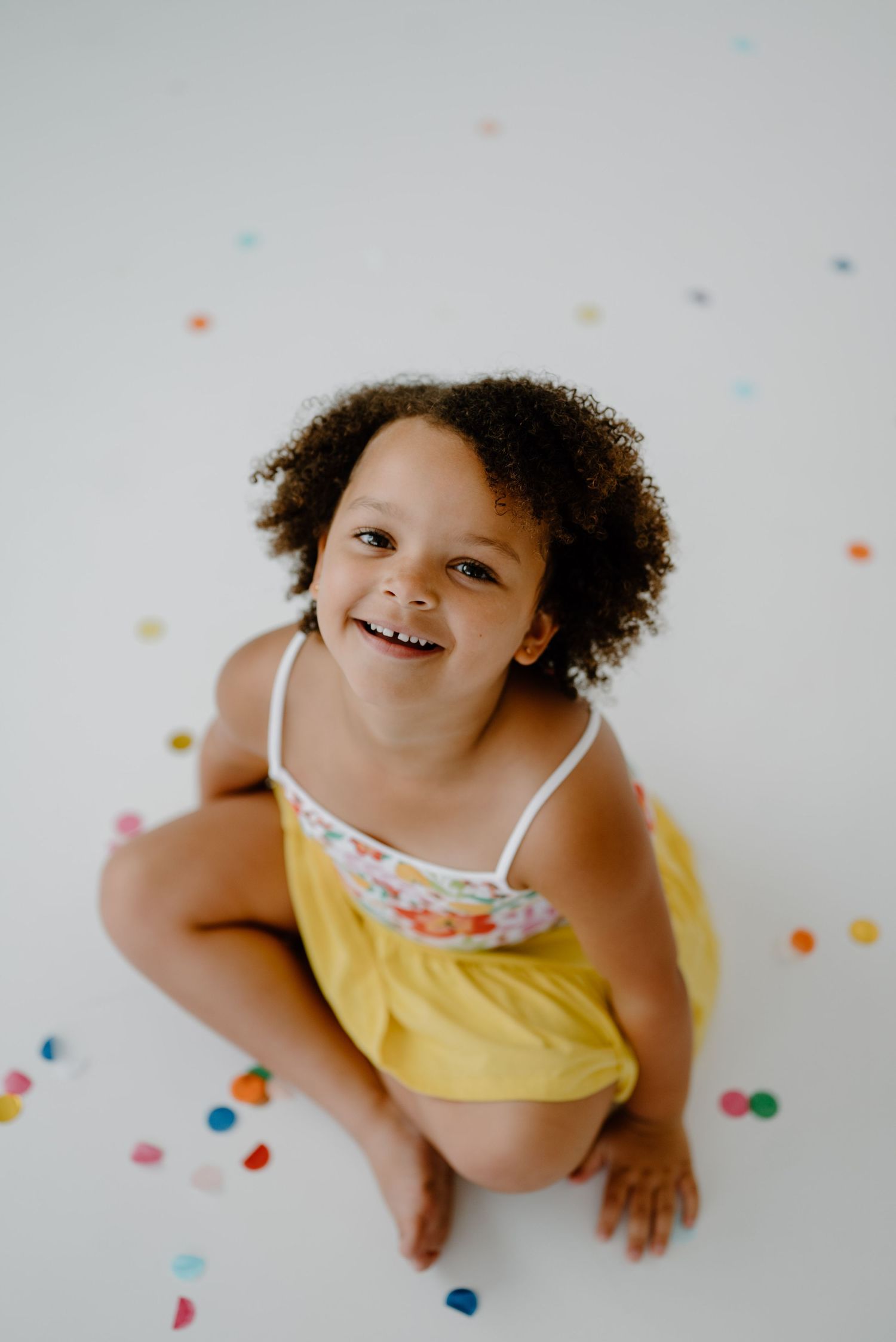 Cheerful toddler girl sitting on floor surrounded by colorful confetti, looking up and smiling.