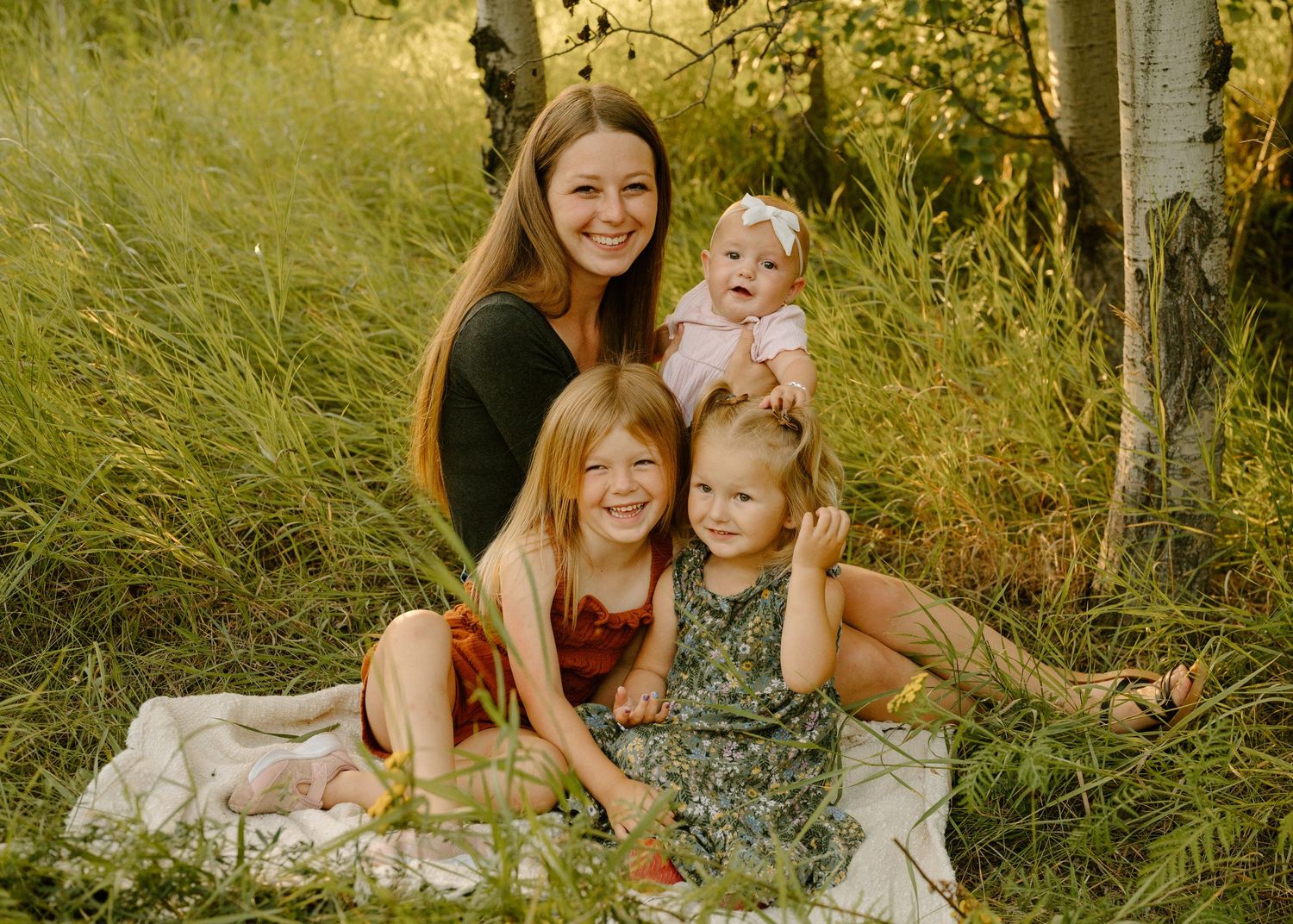 Smiling mother sitting in lush green field with her three young daughters including a baby girl.
