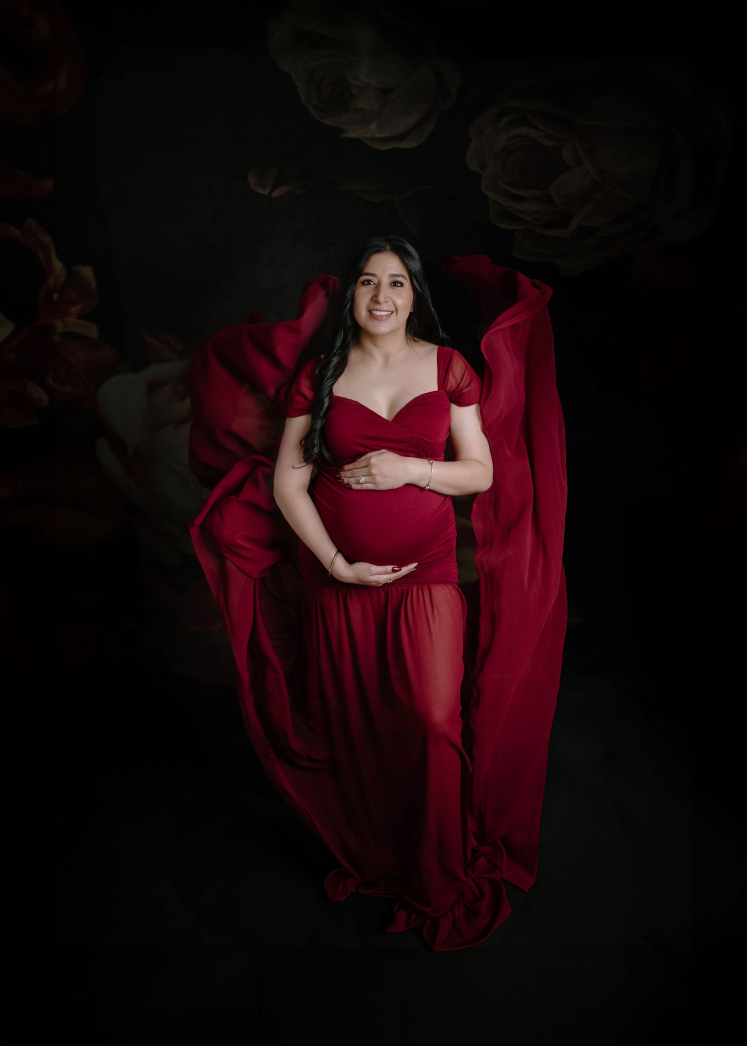 A pregnant woman in a flowing red gown poses with fabric billowing around her in a dark floral studio.