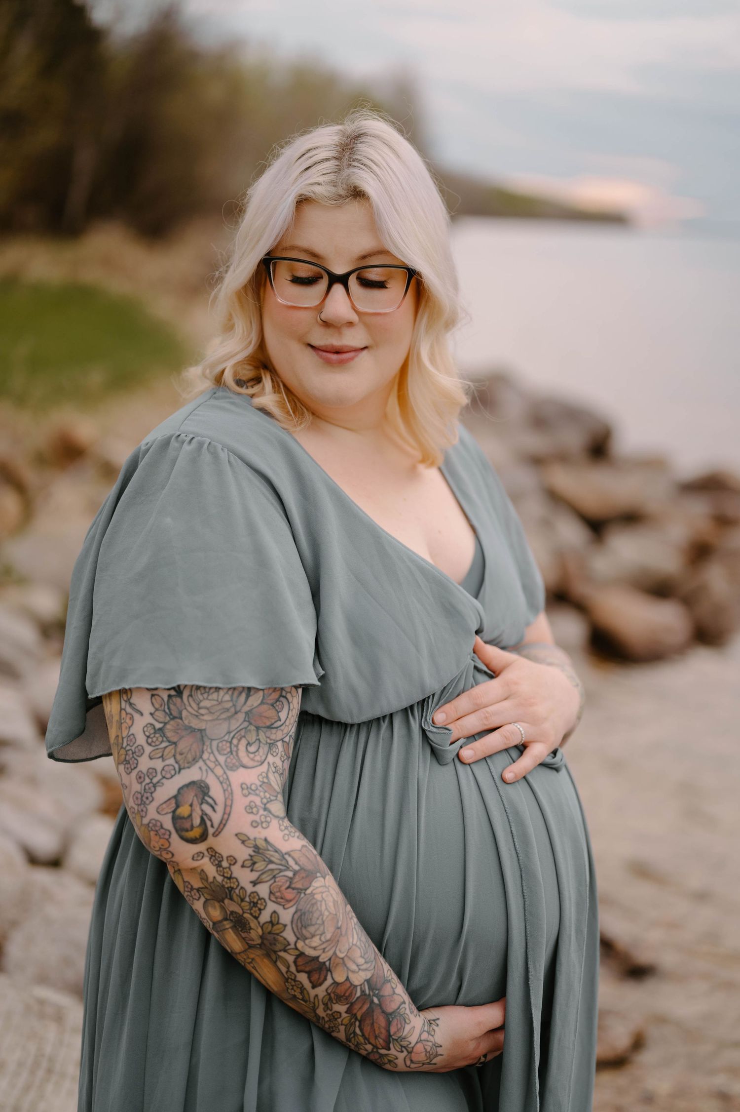 Pregnant woman in sage green dress with tattoo sleeve holds belly near rocky shoreline at sunset.