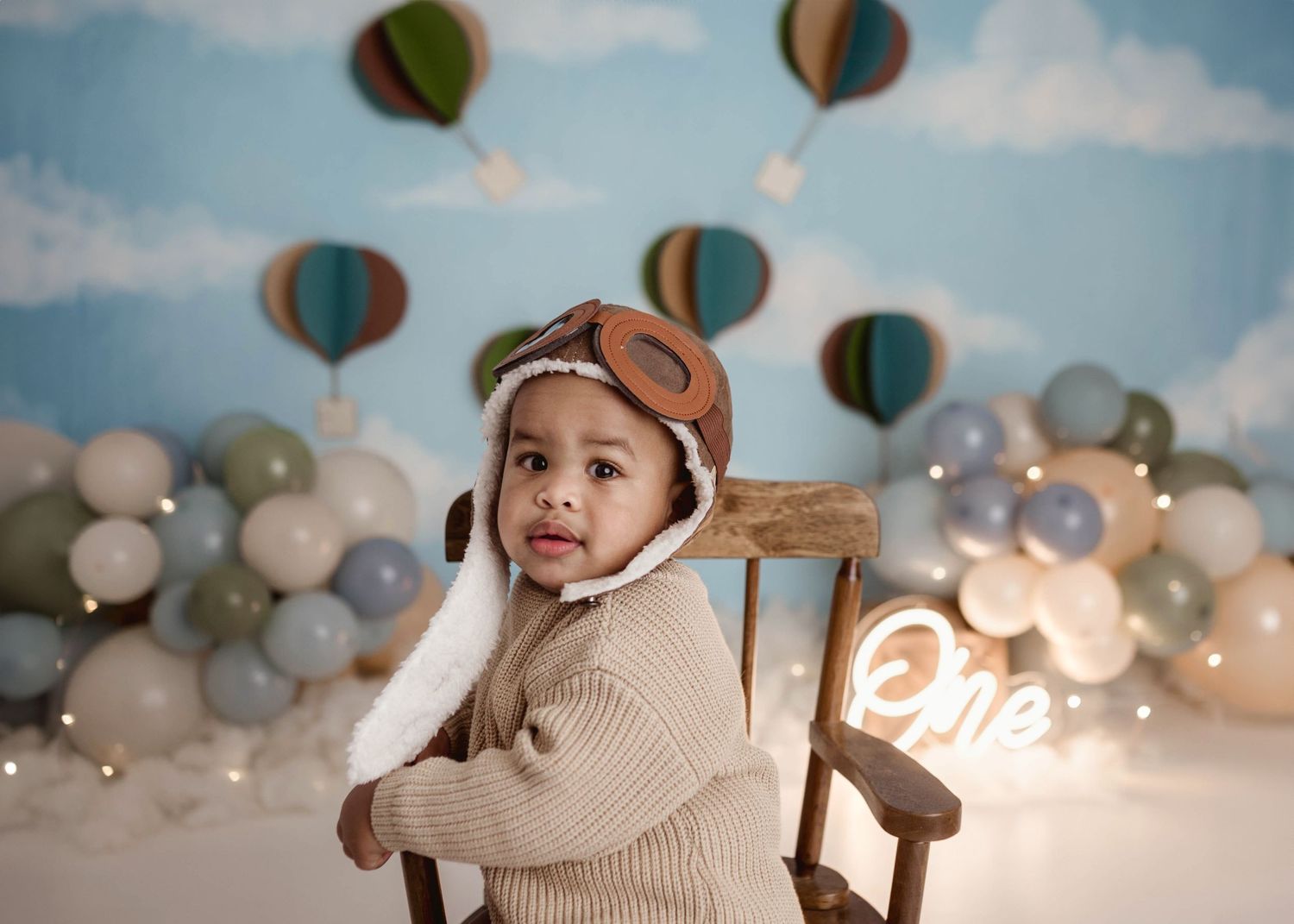 Cute baby in aviator hat standing at a wooden chair at a hot air balloon first birthday session with neon One sign.