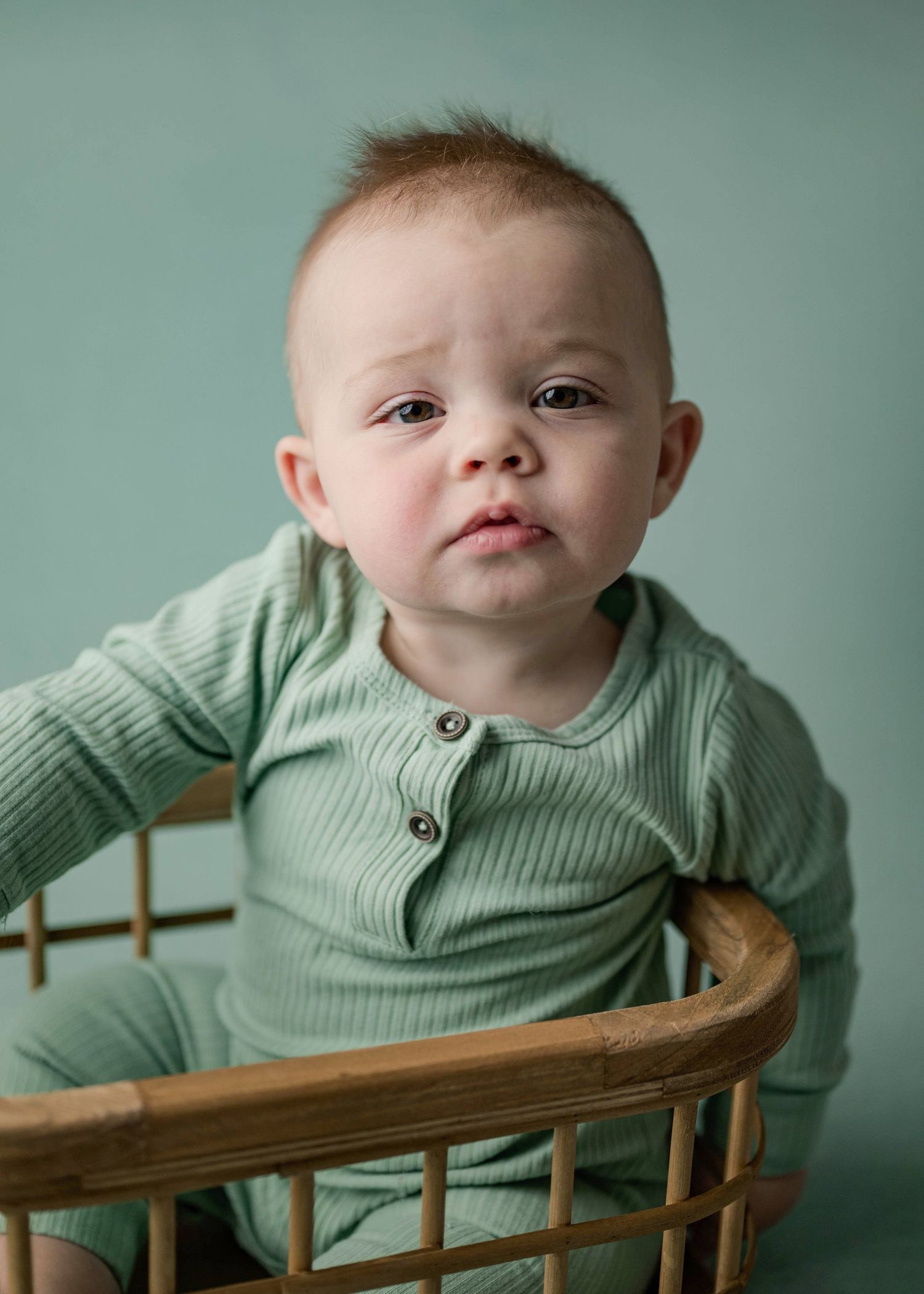 Curious baby boy in green ribbed romper sitting in a wooden wicker basket on teal backdrop.