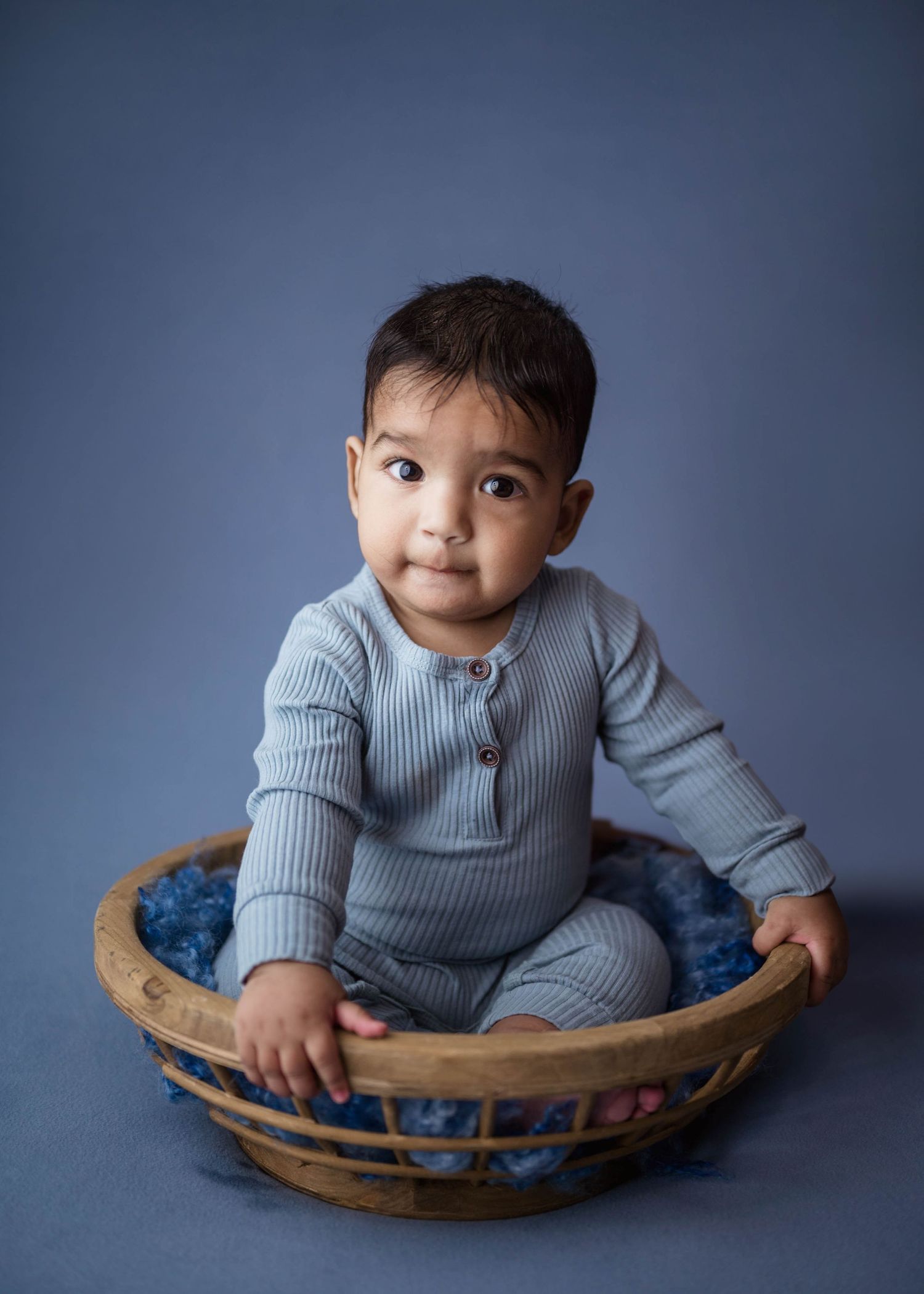Sweet baby in a blue ribbed romper sitting inside a wooden bowl prop on a blue studio background.