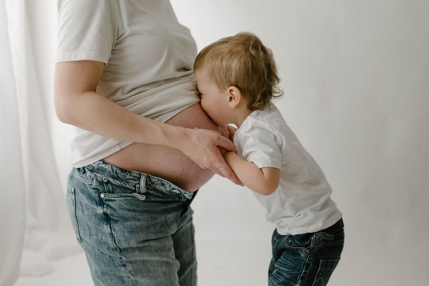 Curious toddler in white tee leans in to kiss pregnant mothers bare belly in bright studio.