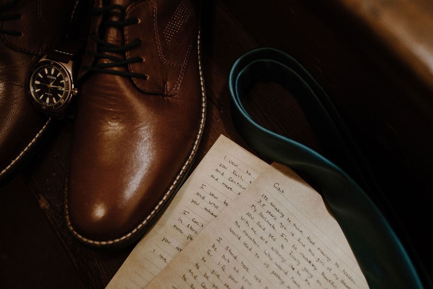 Brown leather dress shoes, watch, teal tie, and handwritten letter arranged as wedding flat lay.