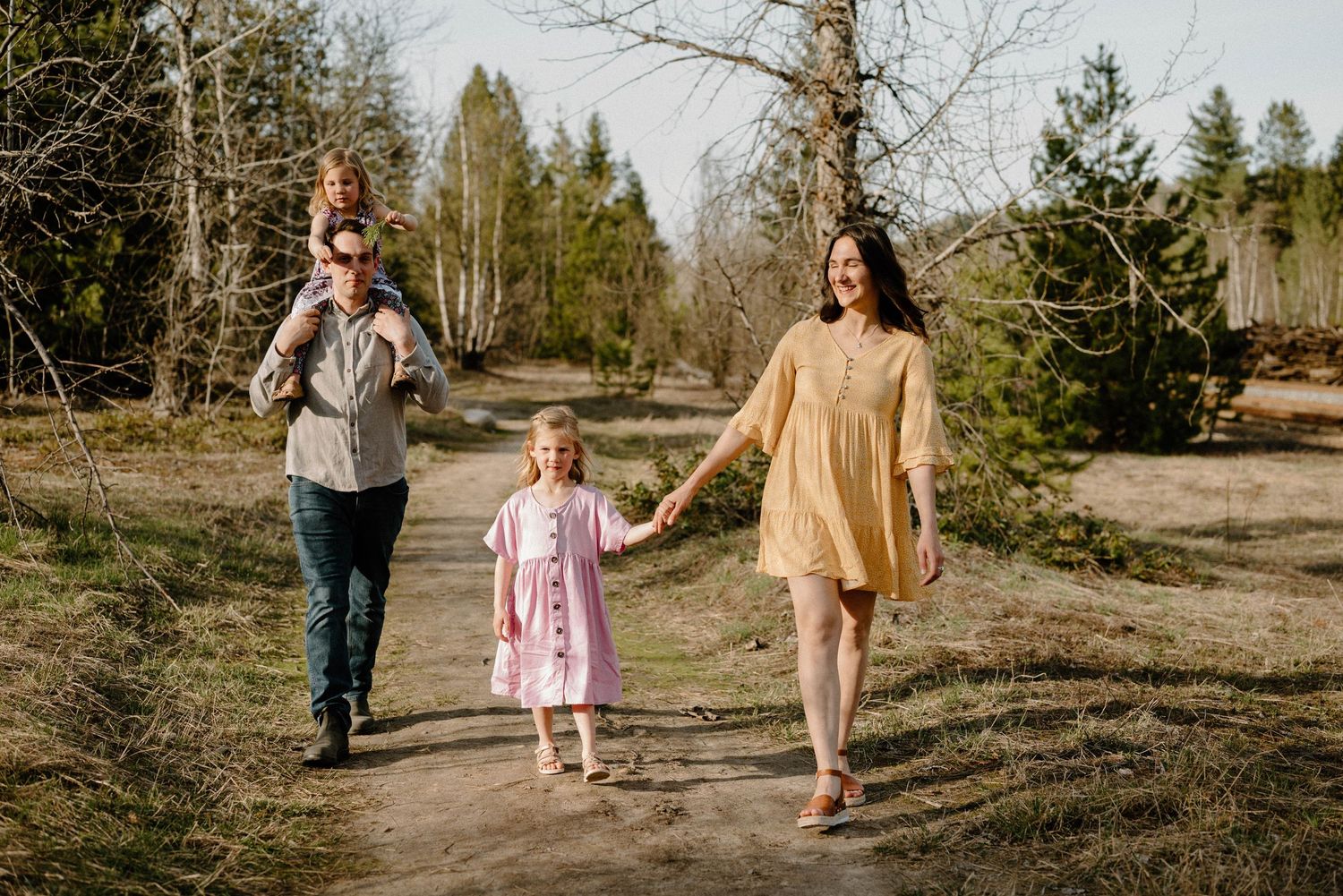 Happy family of four walking hand-in-hand along a sunny forest path in casual outfits.
