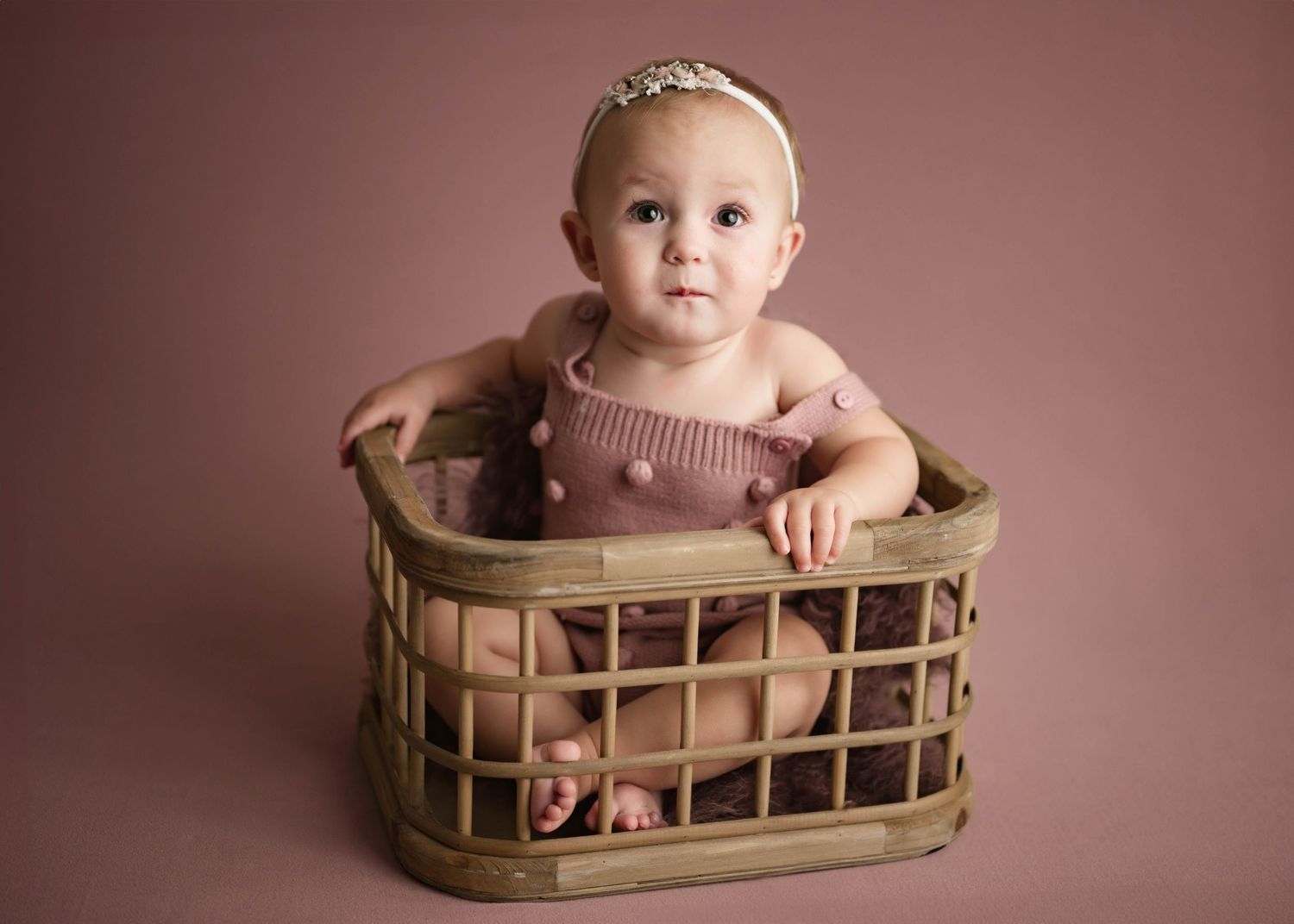 Sweet baby girl in pink knit romper with floral headband posed in rustic wooden basket.