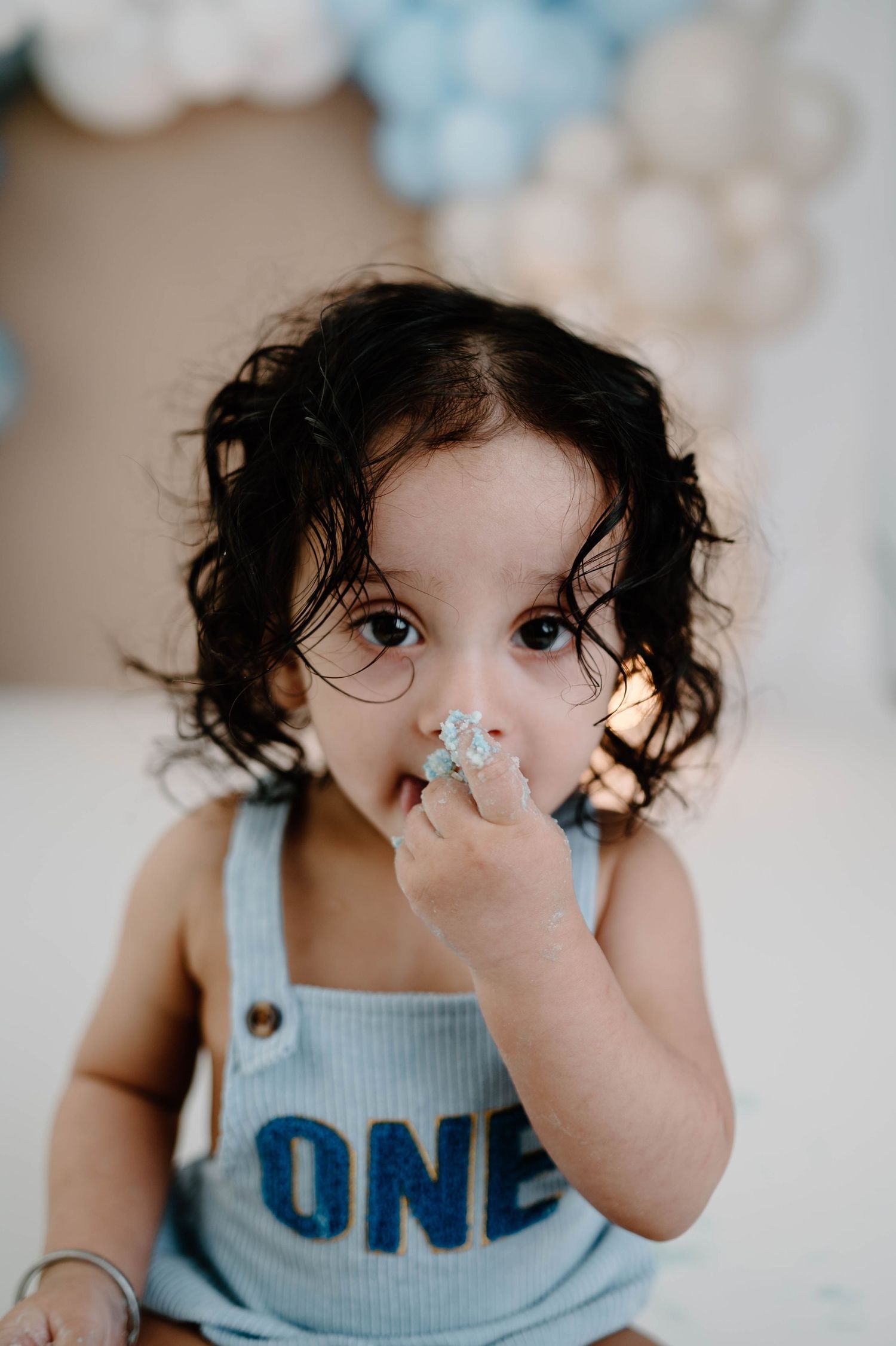 A close-up of a curly-haired toddler in a blue ONE romper eating cake with frosting on their face.