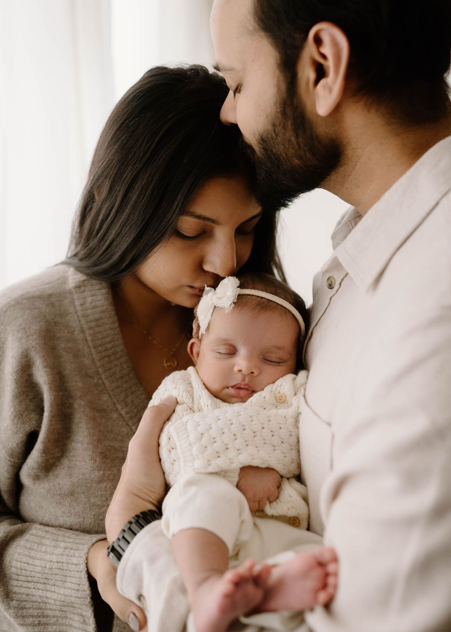 Parents tenderly hold their sleeping newborn baby girl wearing a white bow headband and knit outfit.