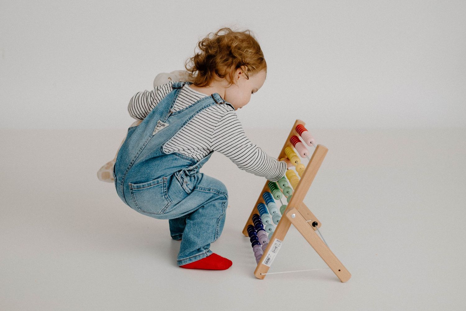 Toddler girl in denim overalls and red shoes crouching to explore a colorful wooden abacus toy.
